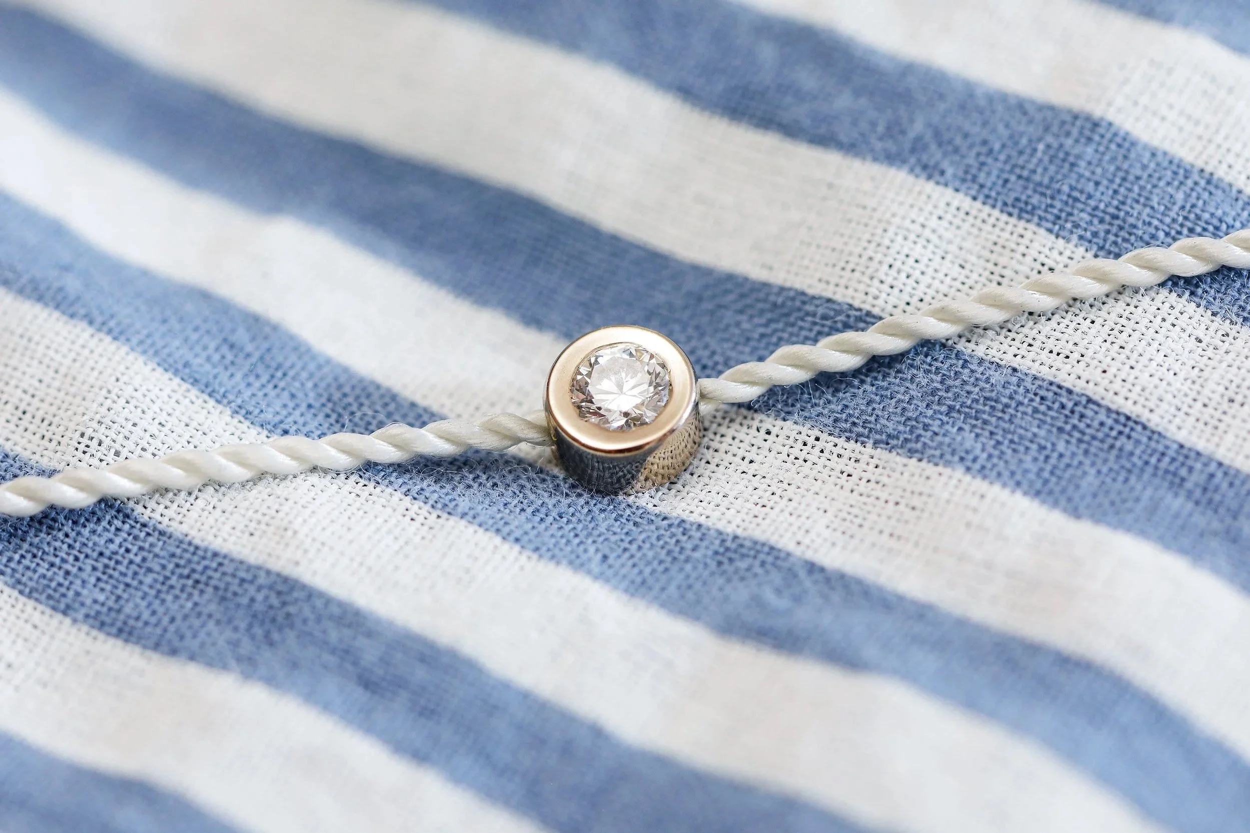 Bracelet avec un diamant de laboratoire serti clos, fixé sur un cordon blanc torsadé, posé sur un tissu à rayures bleues et blanches.