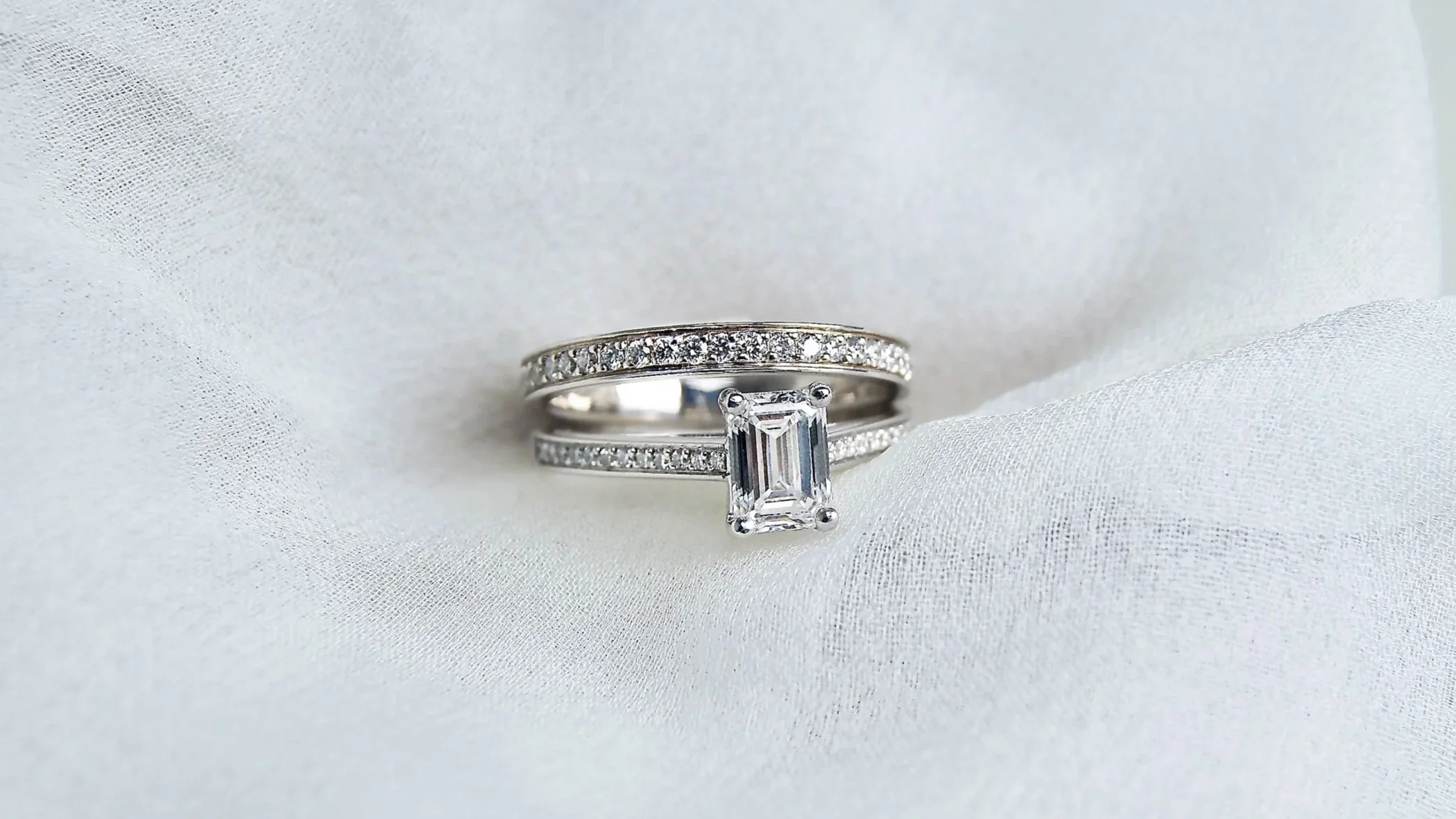 Deux bagues en or blanc l'une est un solitaire avec diamant rectangulaire au centre, et l'autre est une alliance diamant, toutes les deux posées sur un tissu blanc satin.