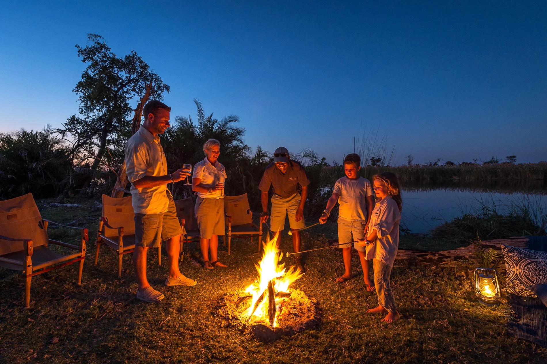 A group of six people, two children and four adults, gathered around a campfire by a river at dusk. They are smiling and enjoying each other's company, some holding drinks and roasting marshmallows. The scene is outdoors with chairs, lanterns, and trees in the background.