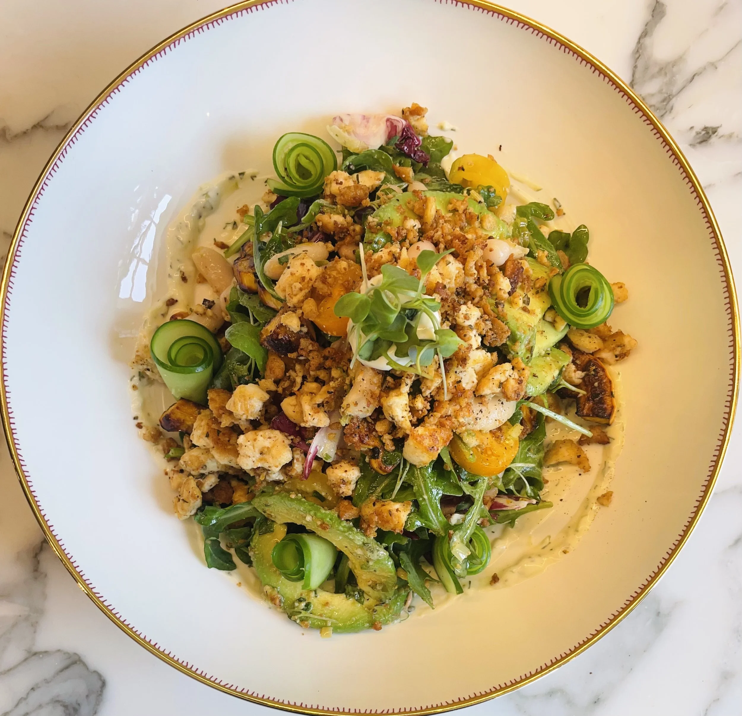 Salad with mixed greens, sliced cucumbers, roasted vegetables, and crumbled tofu, topped with microgreens and served on a bed of arugula, and whipped tahini dressing.