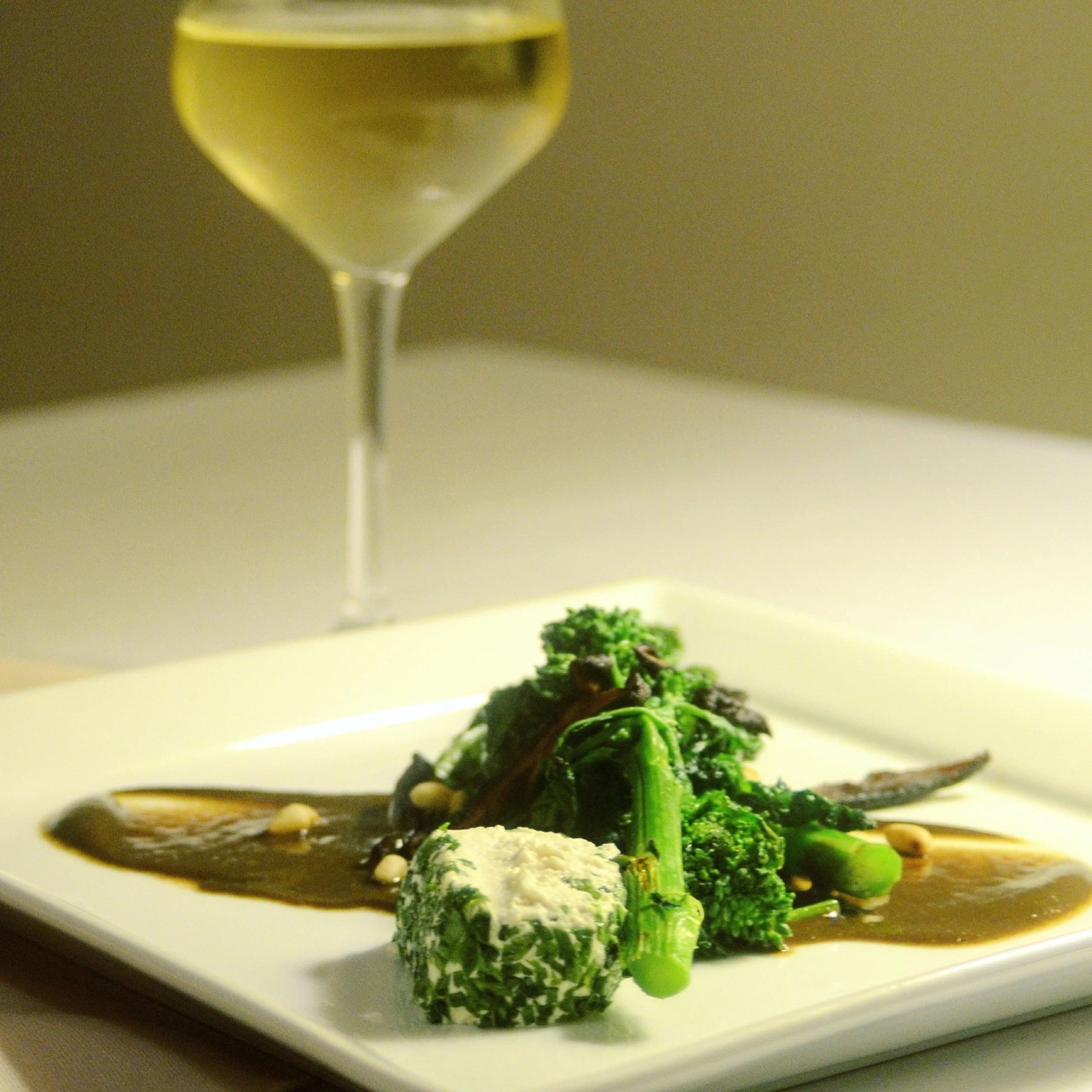 A plated dish of cooked rapini, macadamia chèvre, drizzled with black garlic sauce, with a glass of white wine in the background.
