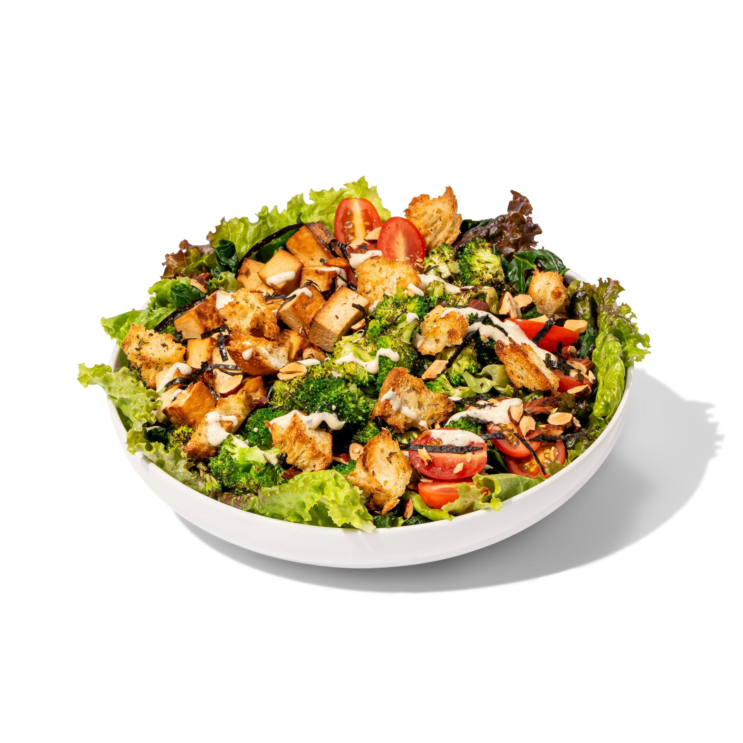 Fresh mixed vegetable salad with cherry tomatoes, broccoli, lettuce, croutons, sliced almonds, smoked tofu, and a creamy caesar dressing in a white bowl