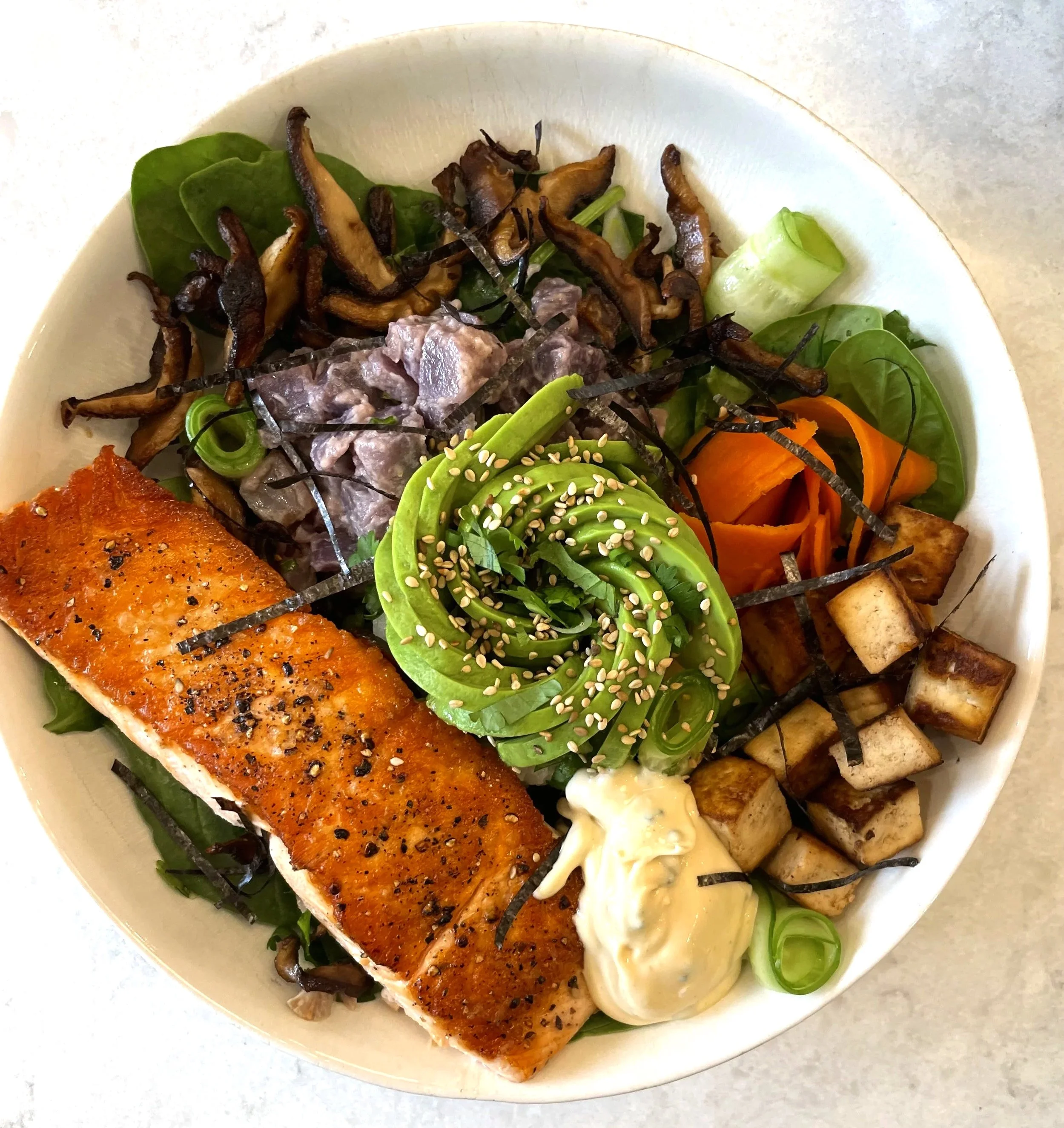 Salmon bowl with greens, avocado, shredded carrots, roasted tofu, mayonnaise, seaweed strips, and shiitake mushrooms.