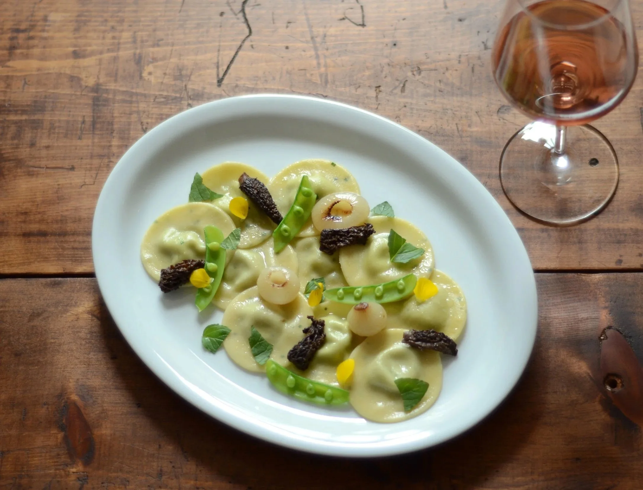 Plate of ravioli pasta garnished with green peas, pearl onions, morel mushrooms, mint, and yellow edible flowers, with a glass of rose wine on a wooden table.