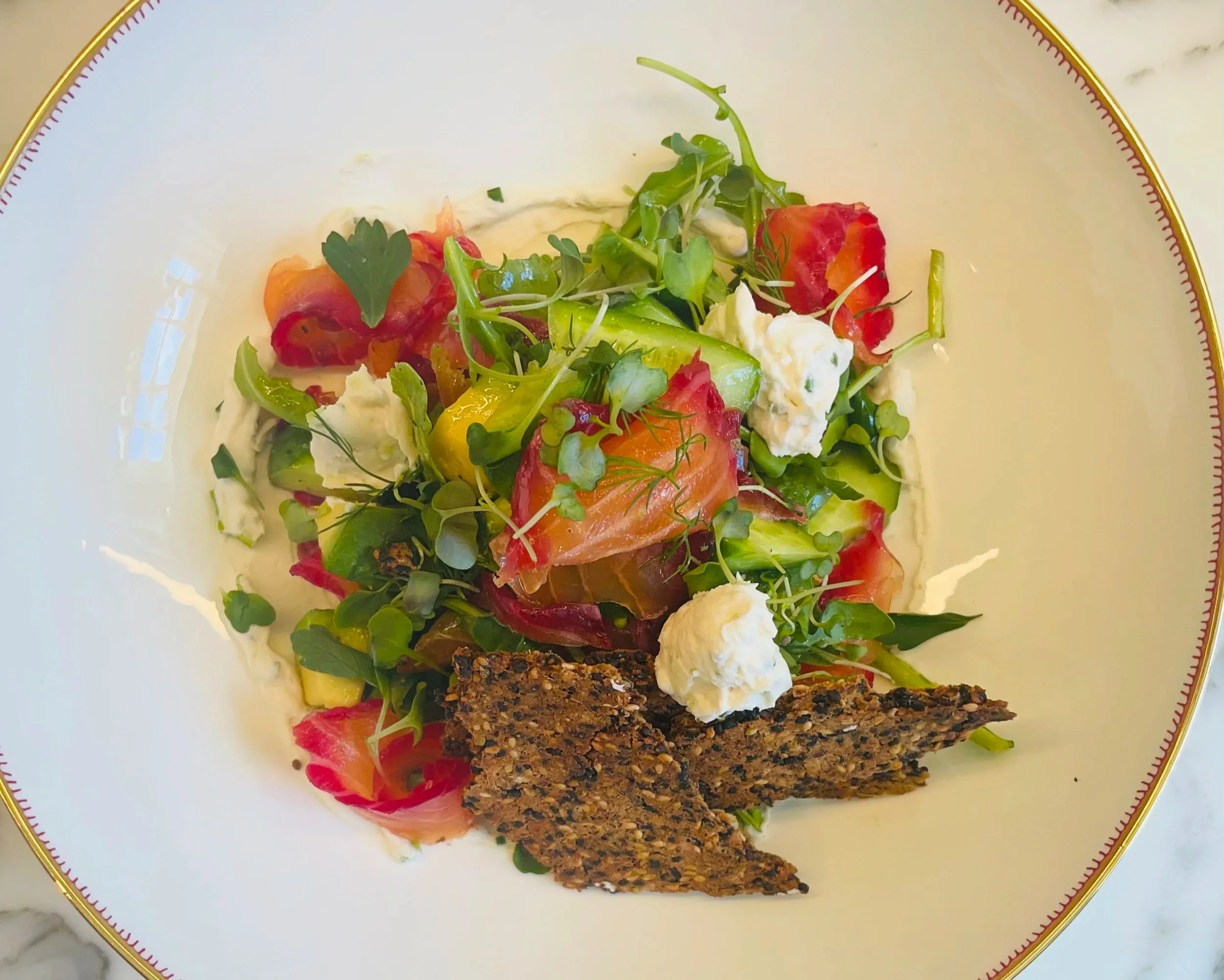 A plate of a gourmet salad with mixed greens, slices of beet cured smoked salmon, crumbled cashew cheese, crispy flax crackers, and fresh herbs, served on a white plate with a gold rim.