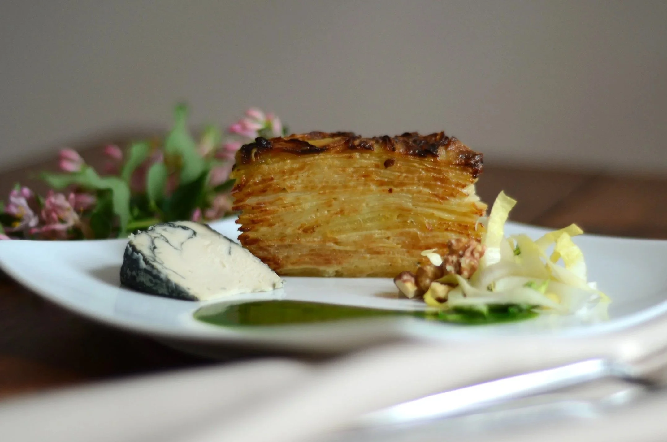 A plate of food featuring a large serving of layered potato and celeriac gratin, a wedge of cashew blue cheese, a small endive salad, and a drizzle of charred scallion oil, with pink flowers in the background.
