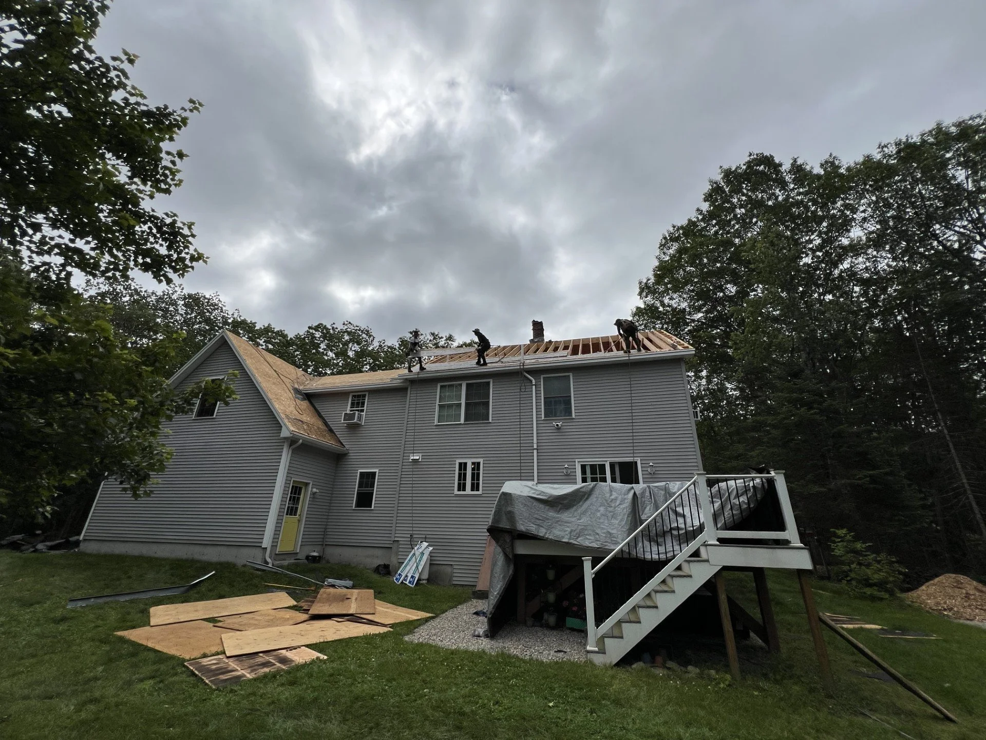 House under construction with workers on the roof, plywood sheets on the ground, and a staircase leading to a deck