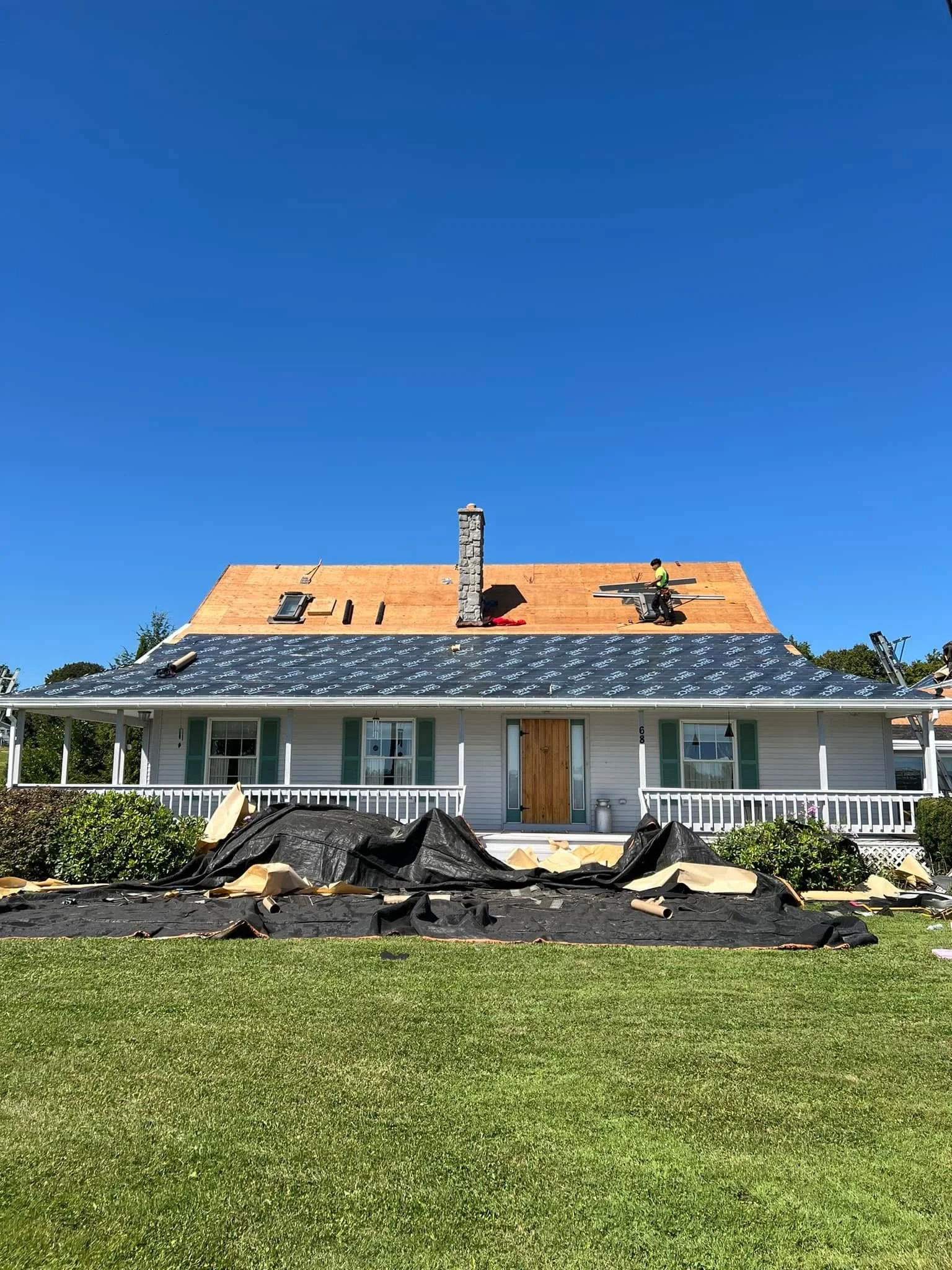 House under construction with roofing work in progress, roofing materials and workers on the roof, and a well-maintained front yard.