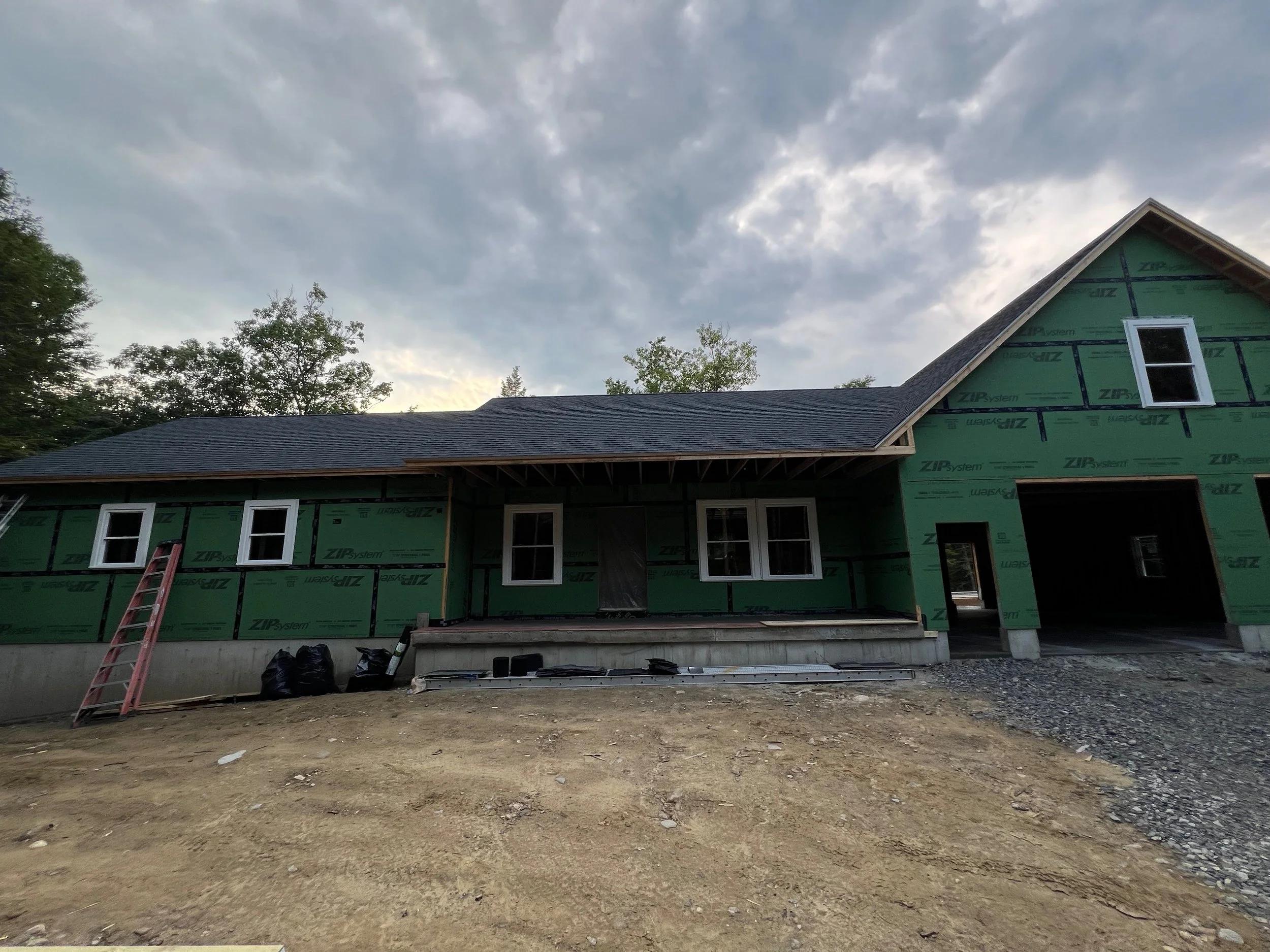 A house under construction with green sheathing, four white-framed windows, a pitched roof, and a garage opening, with a ladder and construction materials in front, under a cloudy sky.
