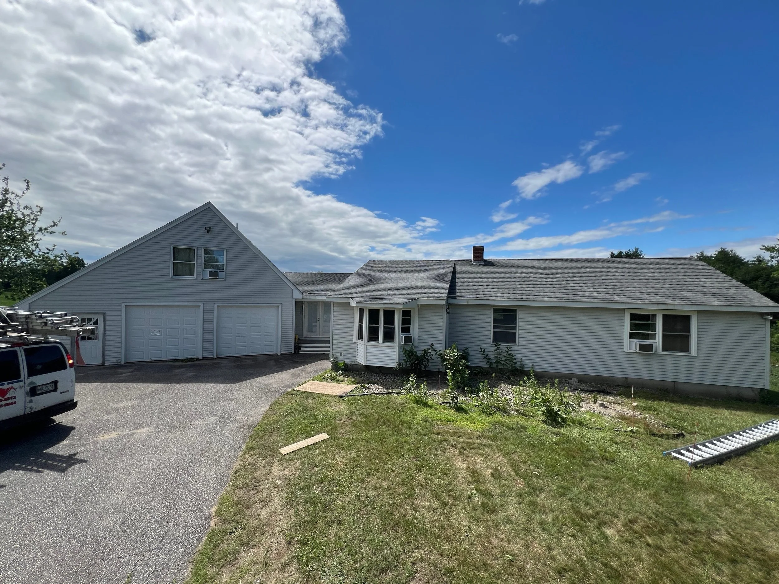 Front view of a residential house with gray siding and a three-car garage, blue sky with clouds, a white van parked to the left, and some plants and a ladder on the yard.