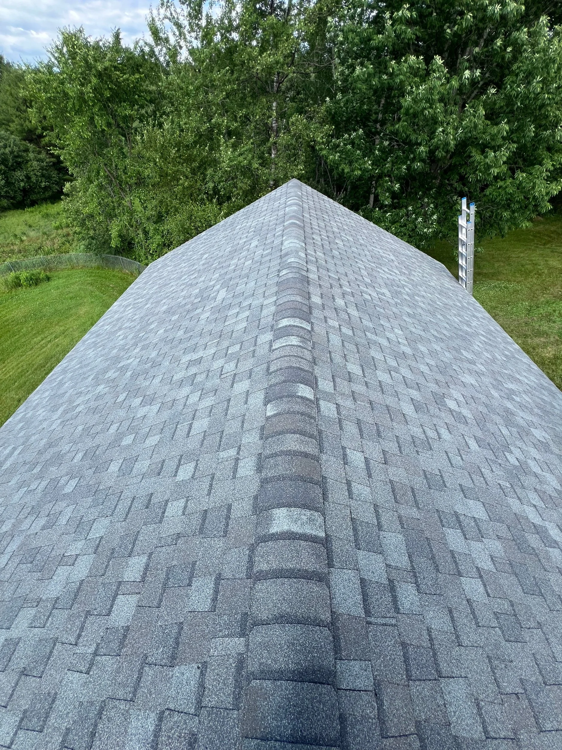 A rooftop in Dayton Maine with asphalt shingles extending into a green landscape with trees and grass, and a ladder on the right side.
