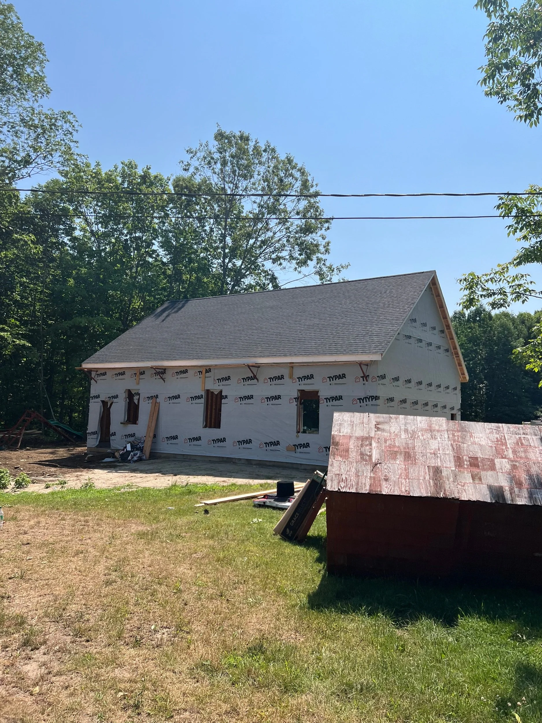 A house under construction with a grey roof and white is wrapped in building wrap, surrounded by green trees and grass, with a red shed in the foreground.