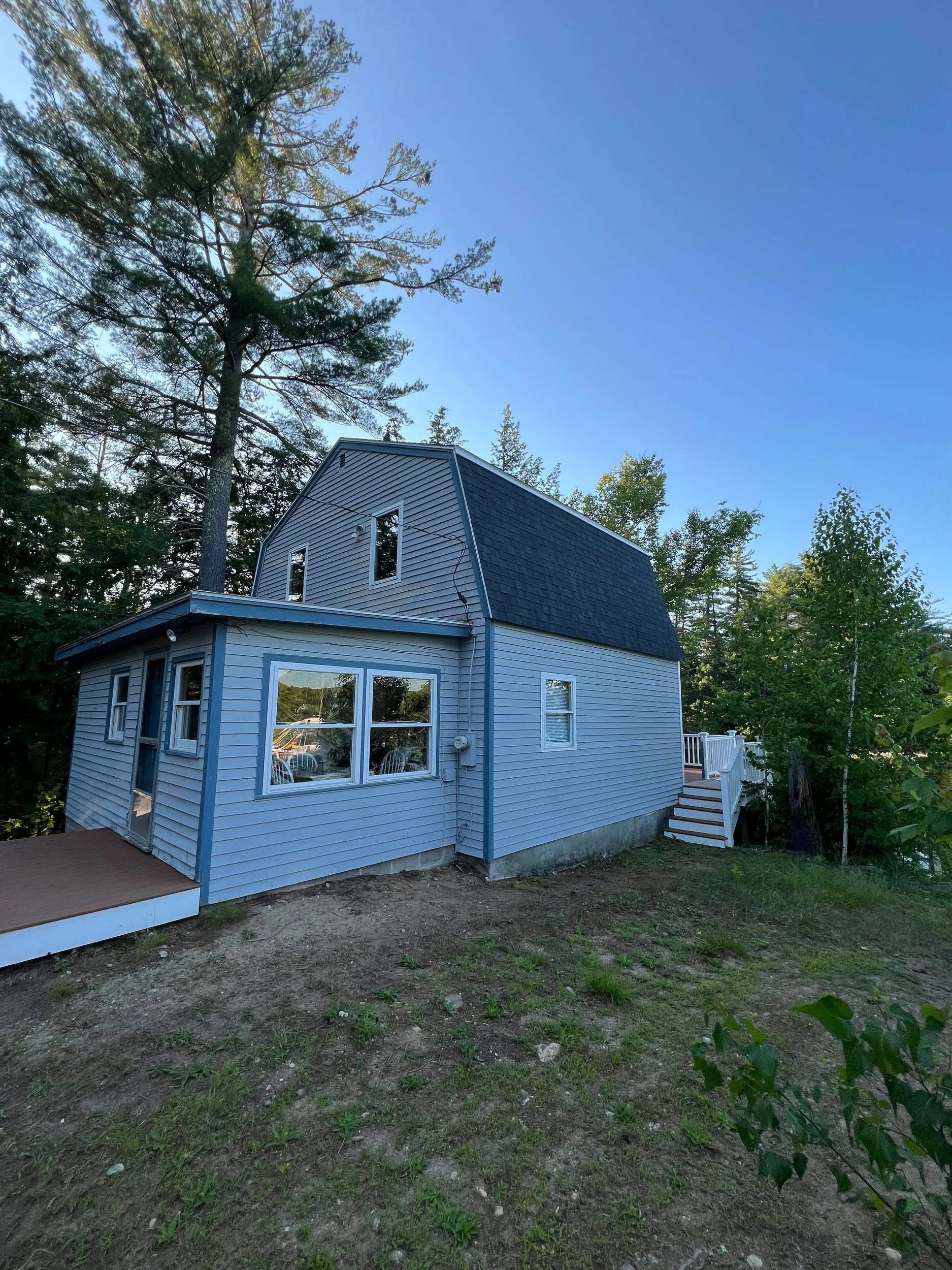 Newly built two-story house painted in light blue with dark blue roof, set in a wooded area with trees around, and a small wooden deck on the side.
