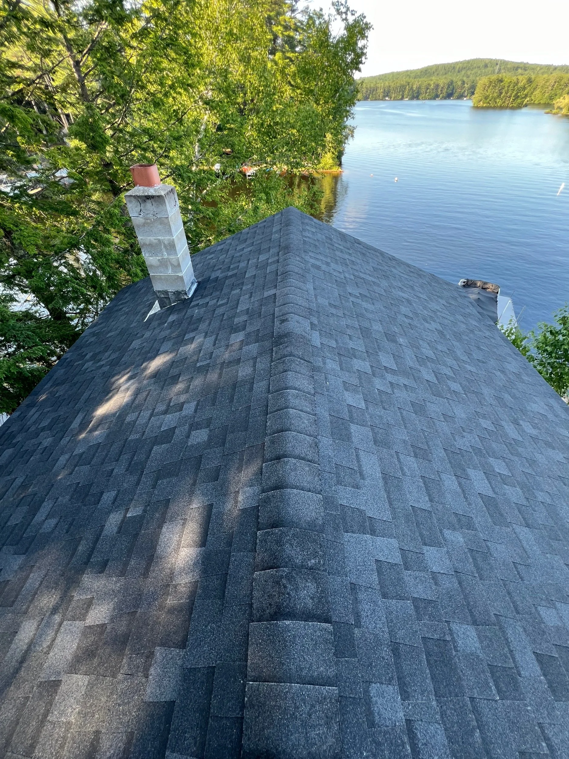 View of a house roof with dark gray shingles, a chimney, and a lake surrounded by trees in the background.