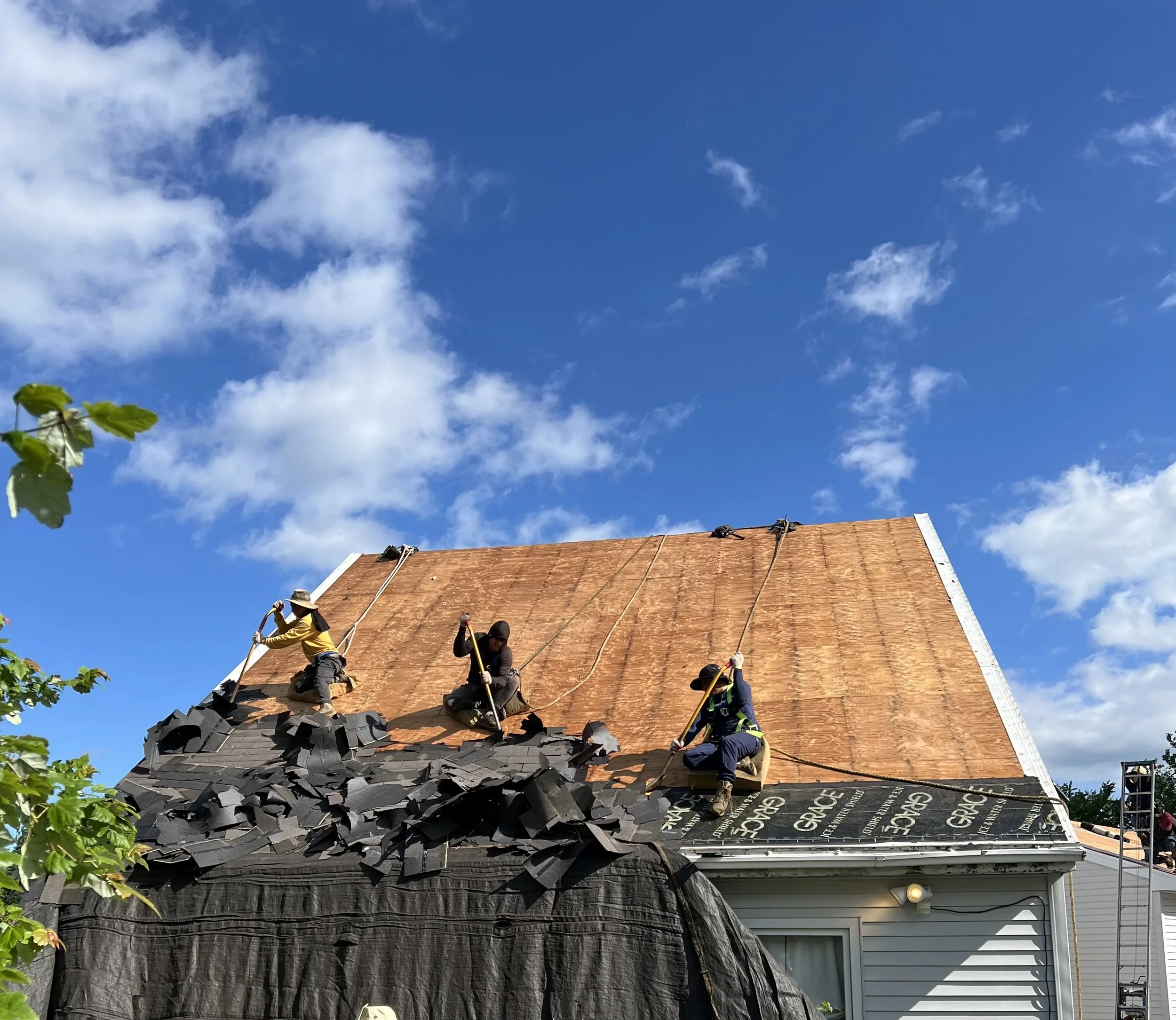 Three workers installing new roofing on a house during daytime, with a blue sky and white clouds in the background.