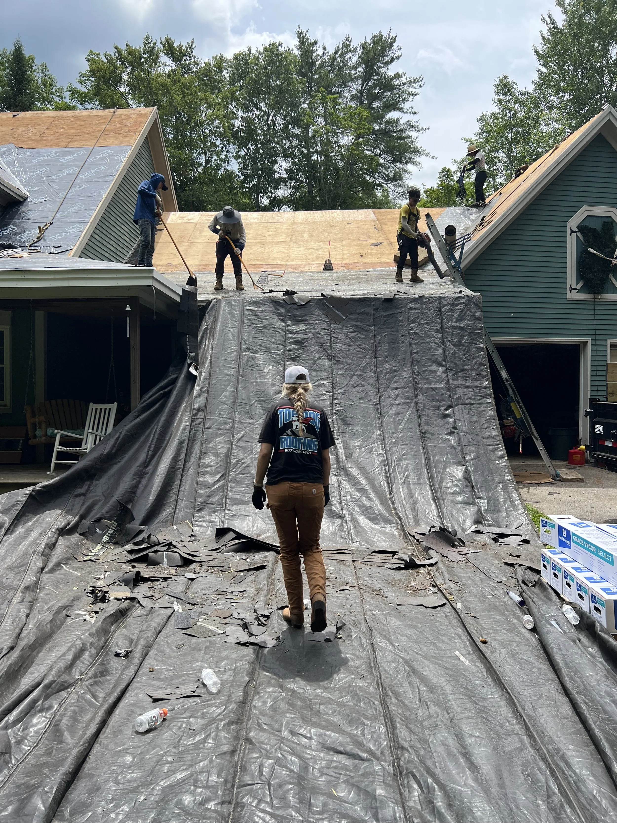 Workers installing a new roof on a house, with some walking on a tarp and others on the shingles, while a woman observes.