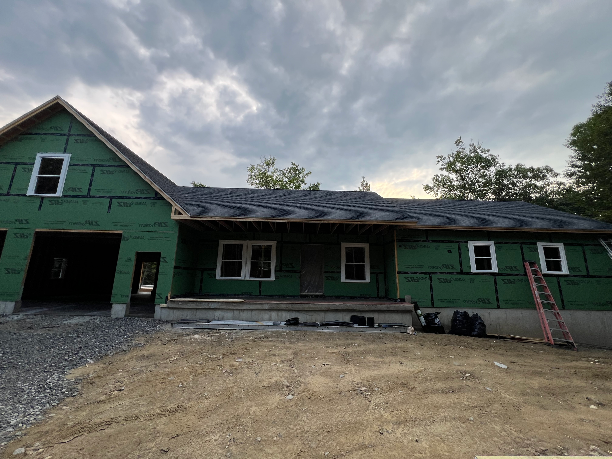A house under construction with green weatherproofing, a black shingled roof, and broken windows in Sanford Maine. The ground in front is bare dirt with construction materials and a ladder leaning against the house.