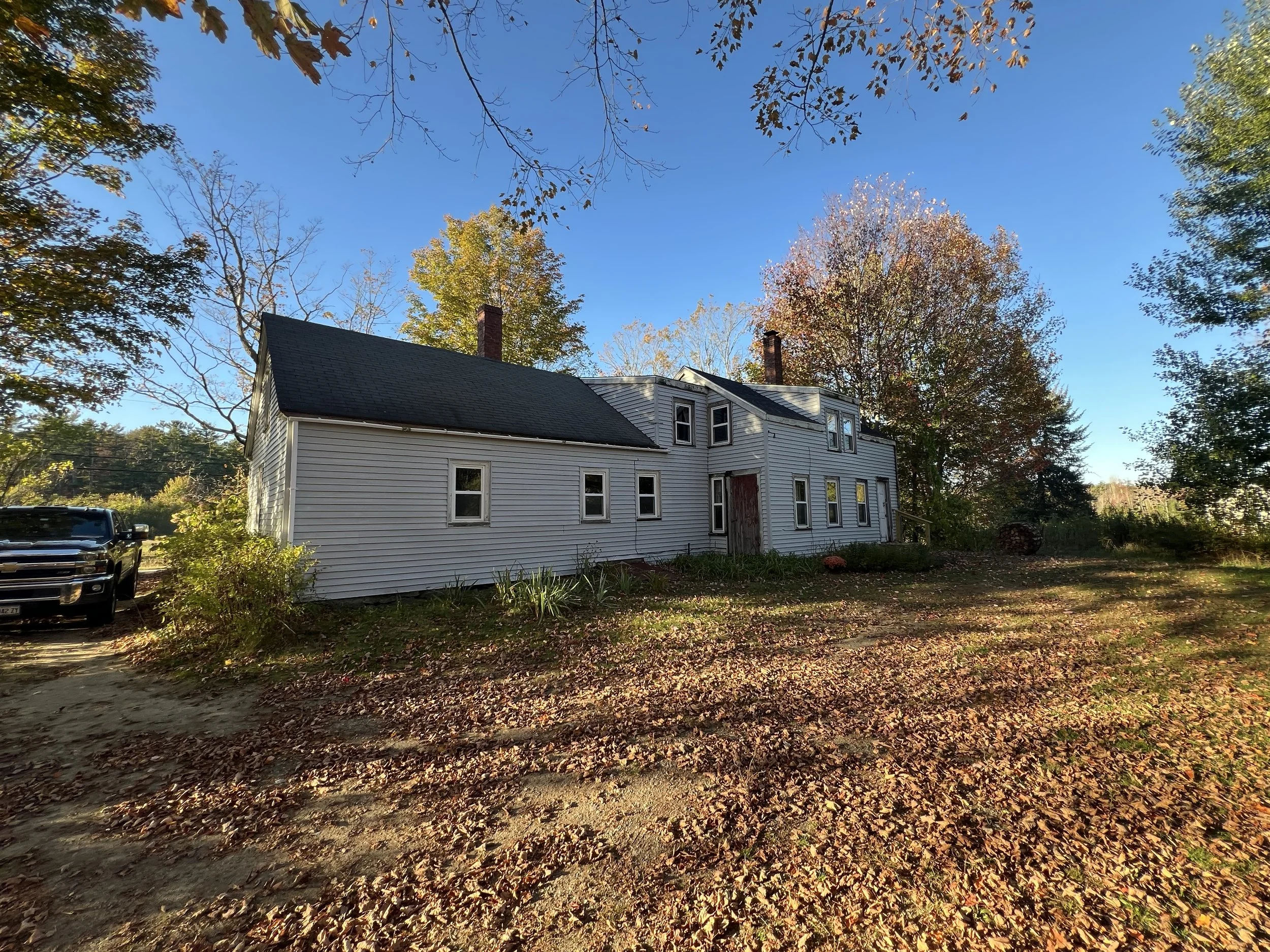 A two-story house with white siding and black roof, surrounded by fall trees with orange and yellow leaves, under a blue sky. A black truck is parked on a dirt driveway to the left.