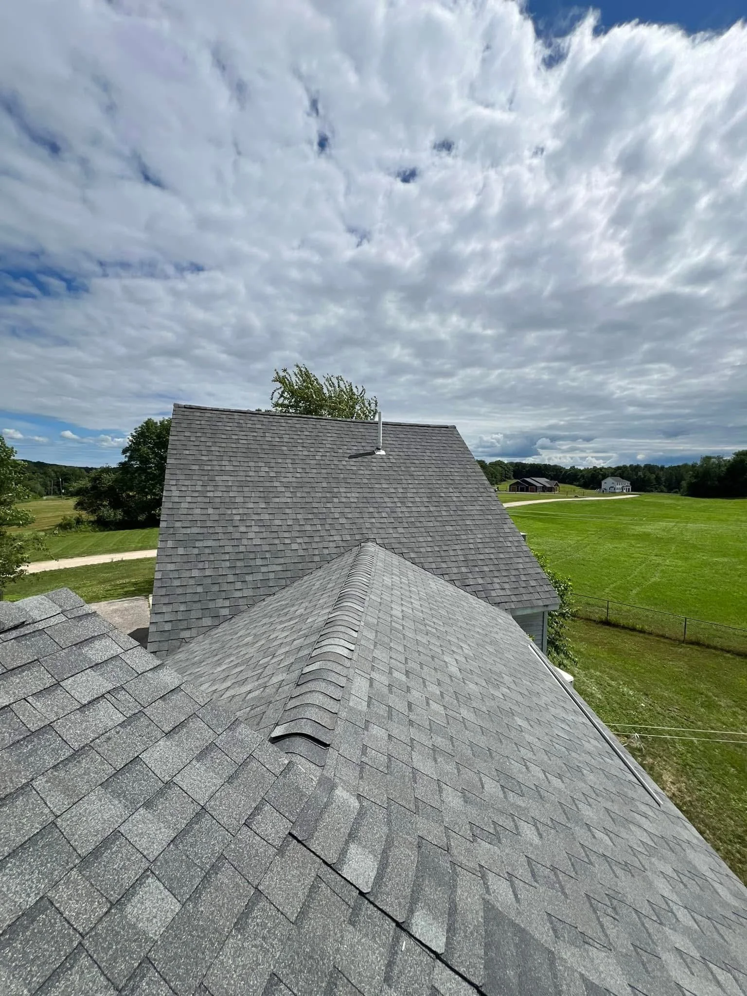 View from rooftops showing gray shingle roofs of two houses, green grass, trees, and a partly cloudy sky.