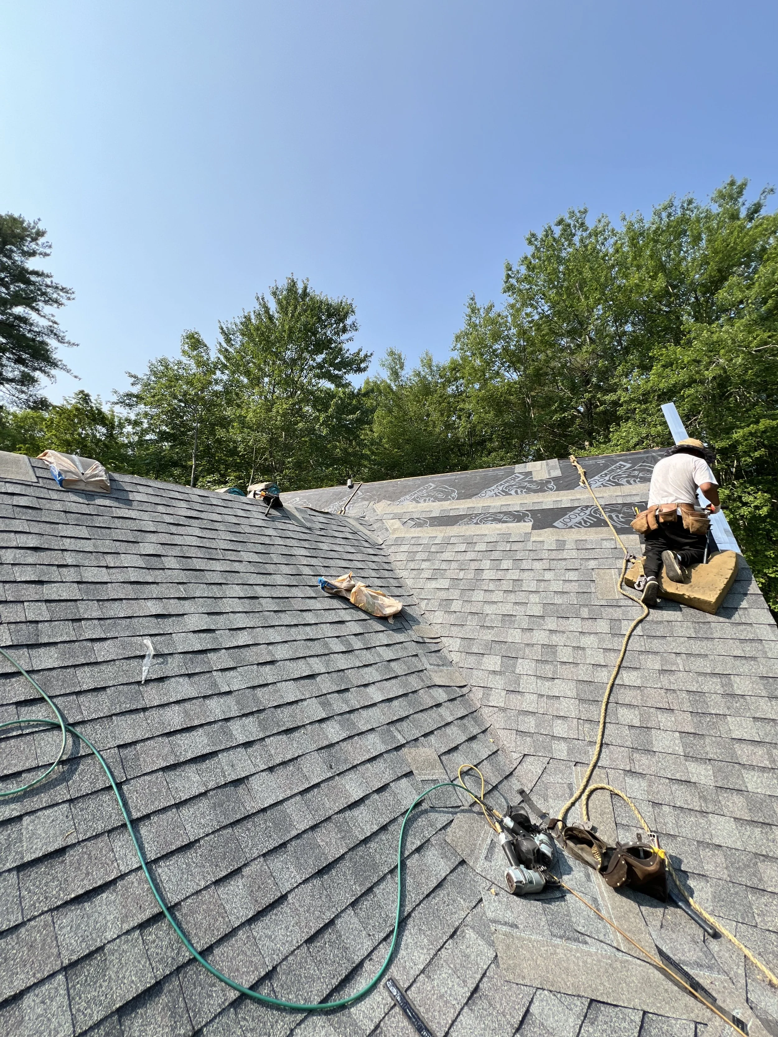 A construction worker on a roof installing roofing materials during daytime, surrounded by trees and a clear blue sky.