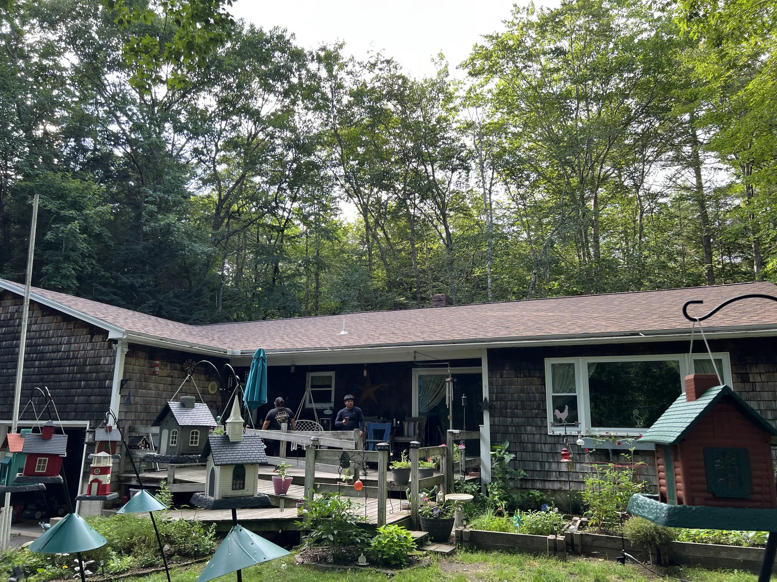 Backyard with garden, birdhouses, patio furniture, and two people standing on the deck, with a wooded forest in the background.