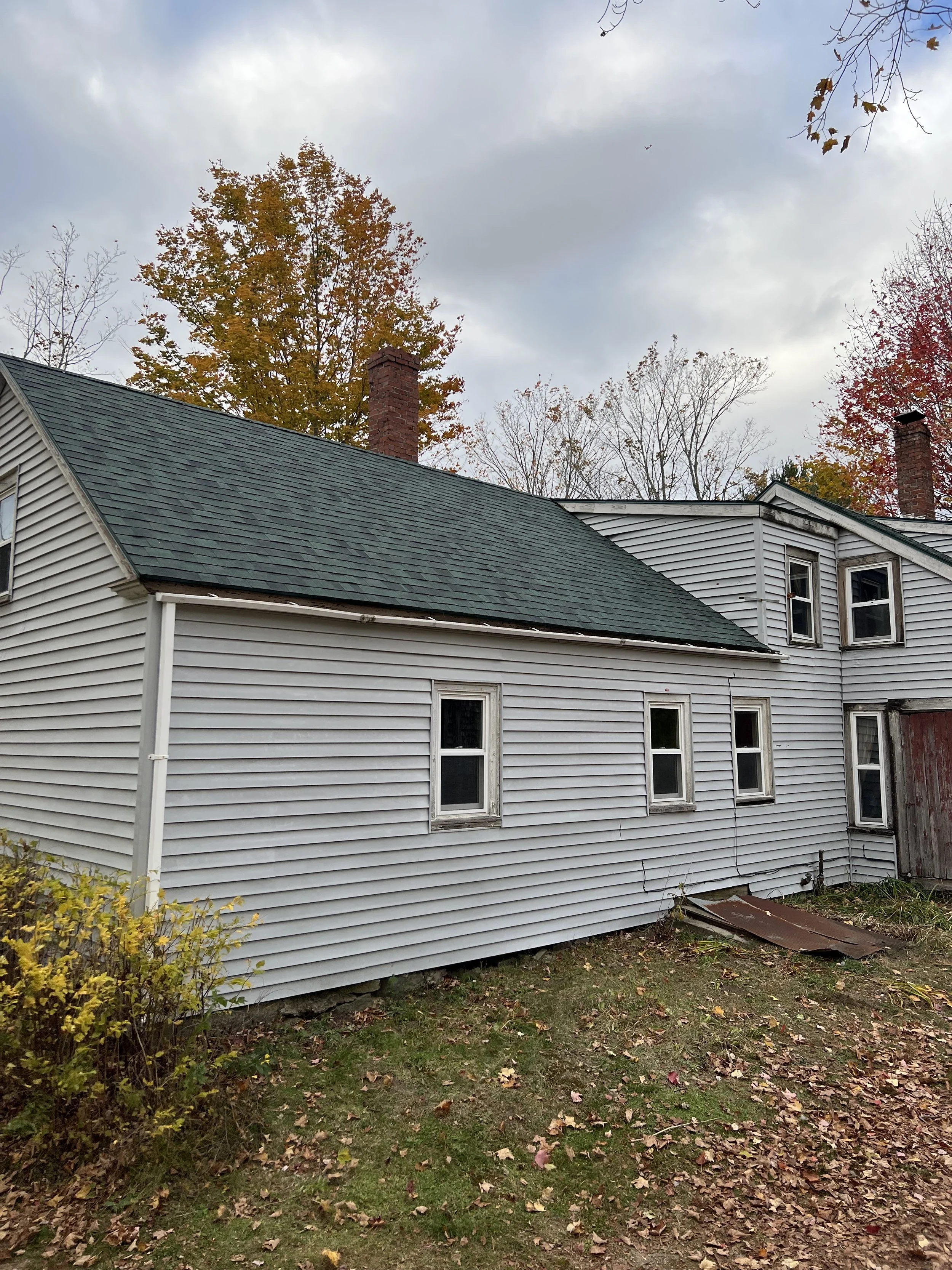 A house with white siding and a dark green roof, several small windows, autumn trees with orange and red leaves in the background, and a cloudy sky overhead.