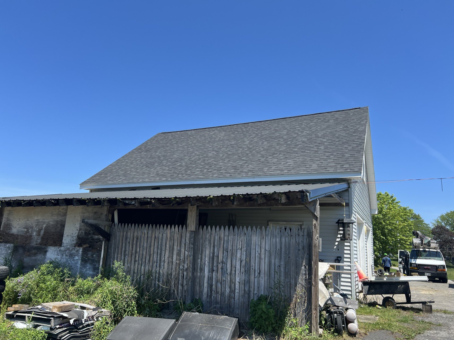 A backyard scene featuring a two-story white house with vinyl siding under a clear blue sky. An adjacent shed with a weathered wooden fence, cluttered with various items like tires, plants, and tools. In the background, a white utility truck and a pe