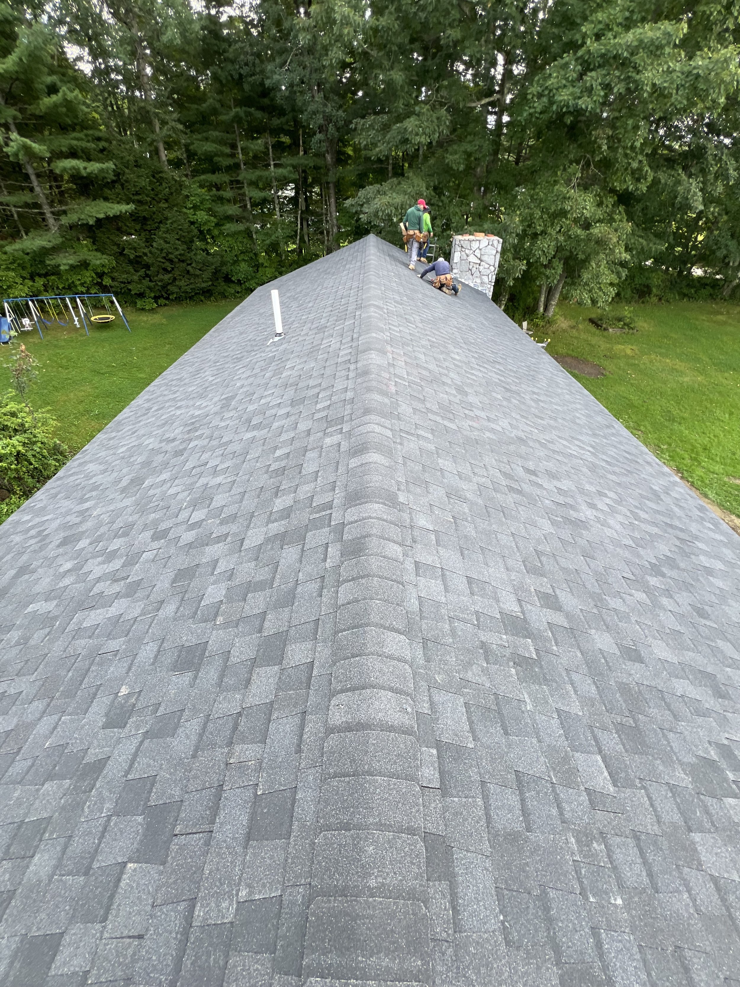View from the rooftop showing four workers installing or repairing shingles on the roof of a house, with a chimney on the side, surrounded by green trees and a backyard with a swing set.