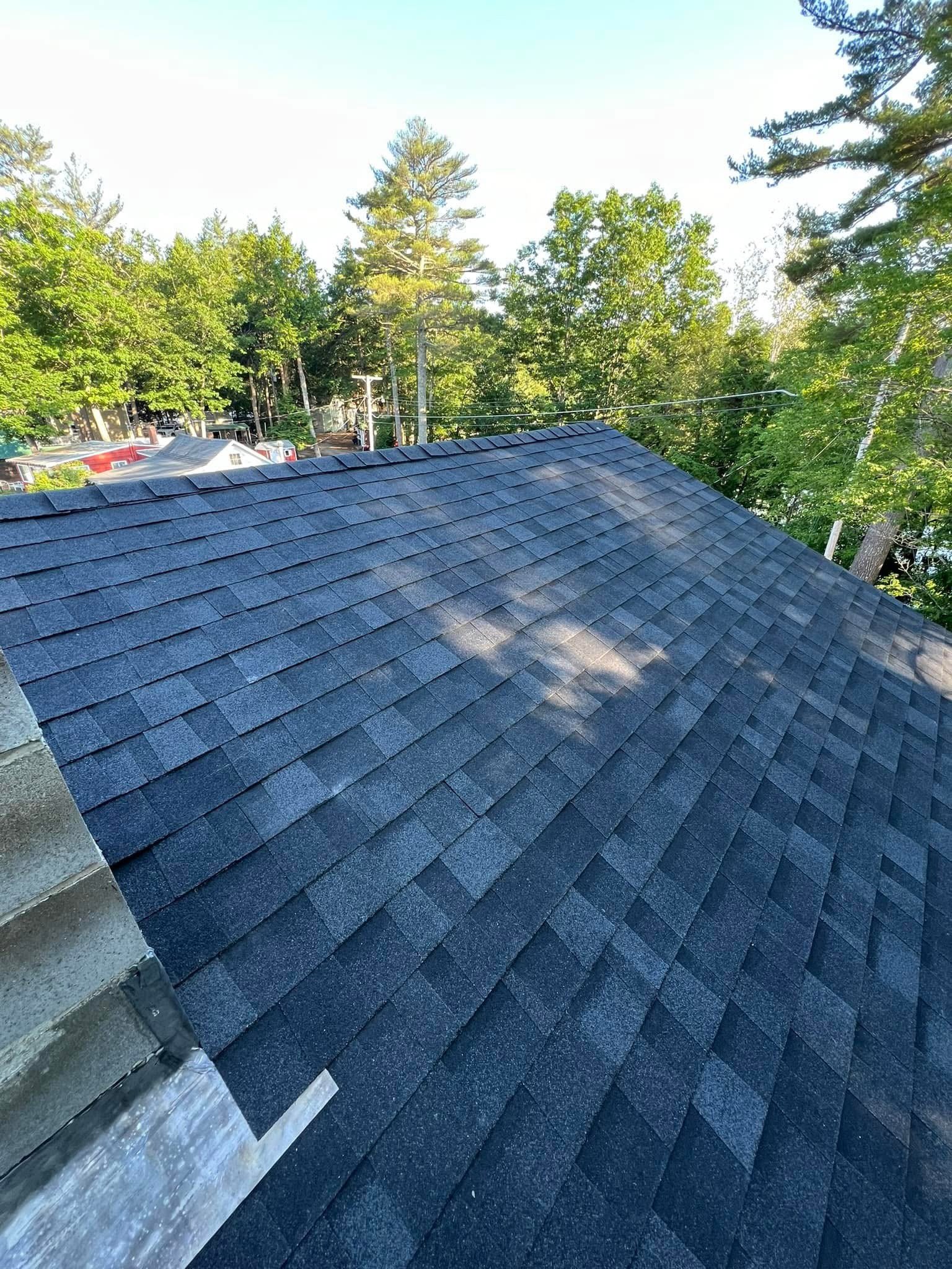 View of a house roof covered with new dark gray asphalt shingles, with surrounding green trees and clear blue sky.