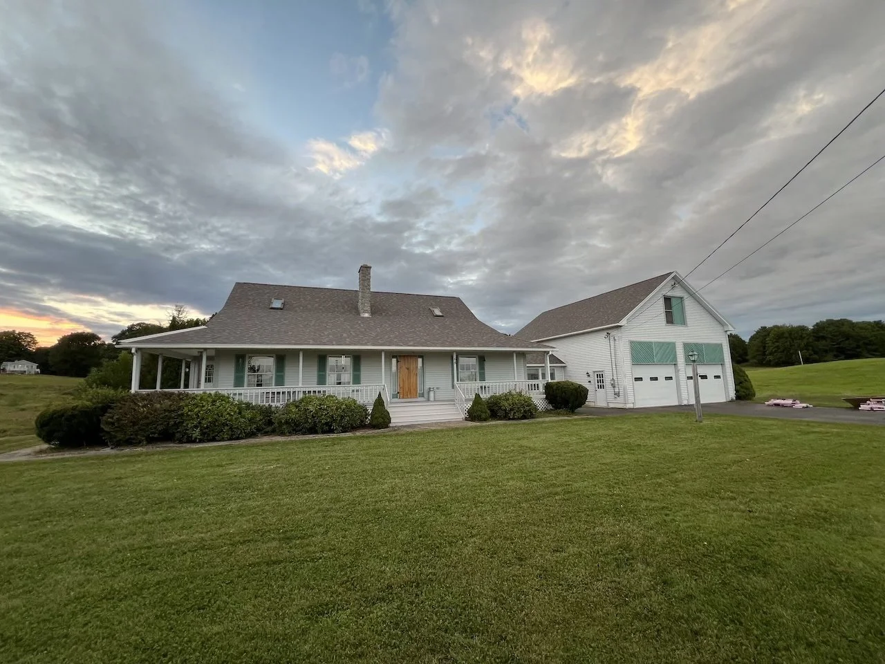 A large suburban house with a porch, steps, and a front yard. The house has a garage and is surrounded by green grass and shrubs. The sky is partly cloudy with the sun setting.