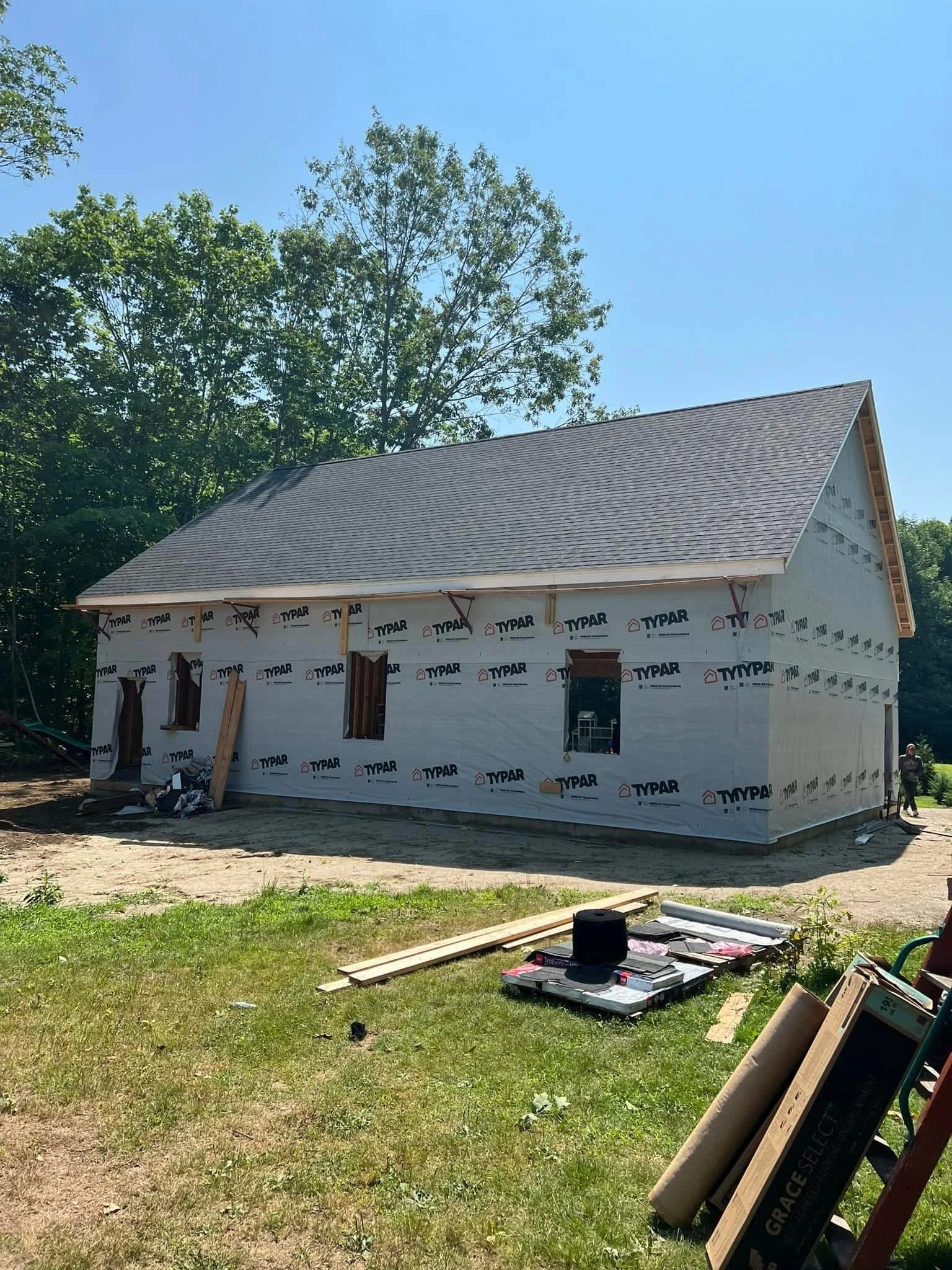 A house under construction with the exterior walls covered in Typar house wrap, surrounded by construction materials and tools, and trees in the background.