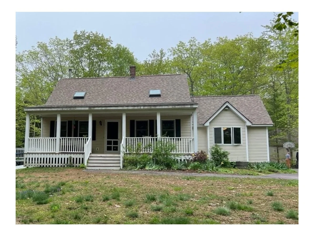 Front view of a two-story house with a porch, white railing, and steps, surrounded by a yard with patches of grass and bushes, with tall trees in the background, and a basketball hoop on the right side.