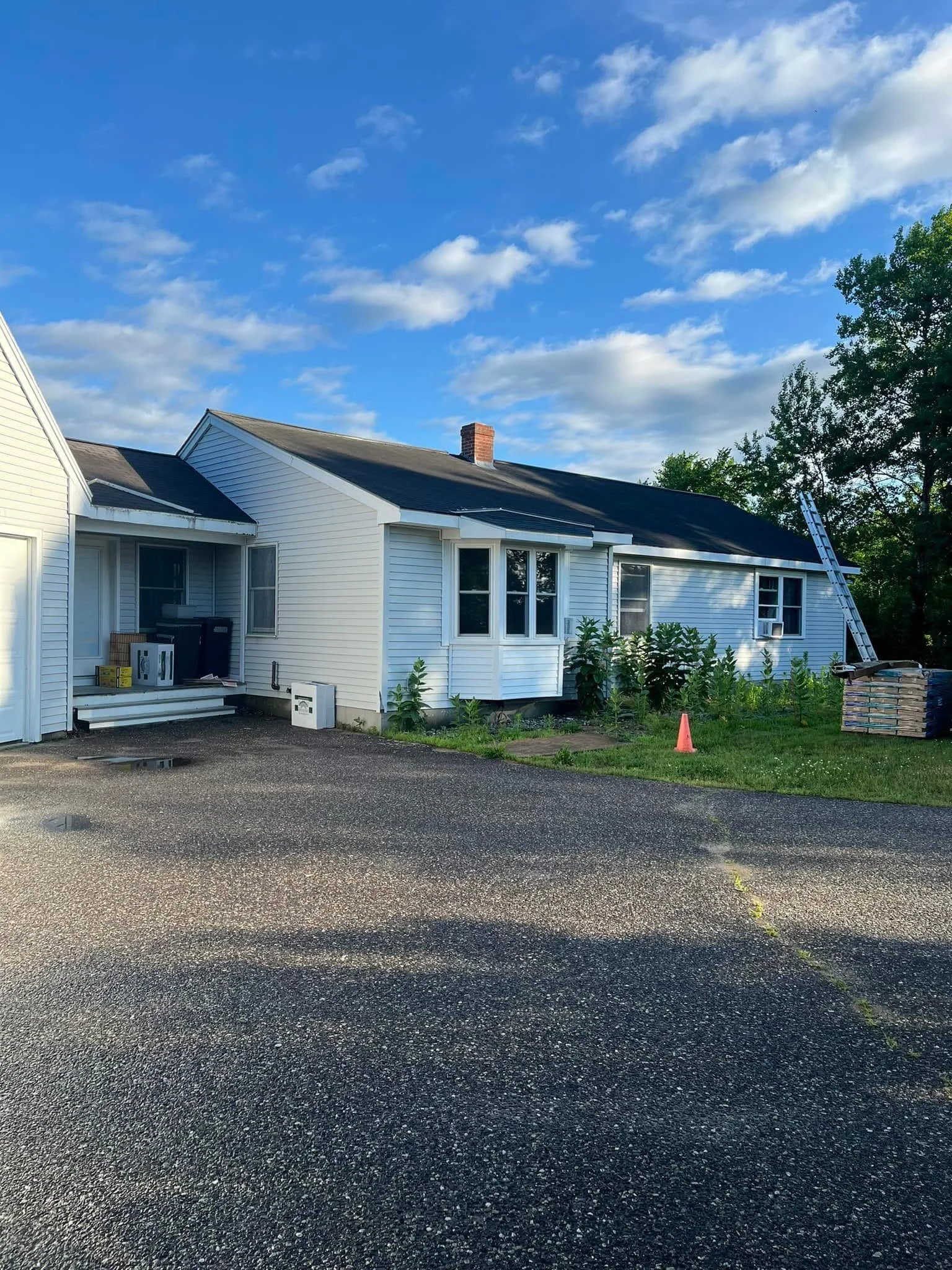 A white house with a dark roof and chimney under a blue sky with scattered clouds. There is a driveway in front and some greenery along the side of the house. A ladder is leaning against a tree on the right side.