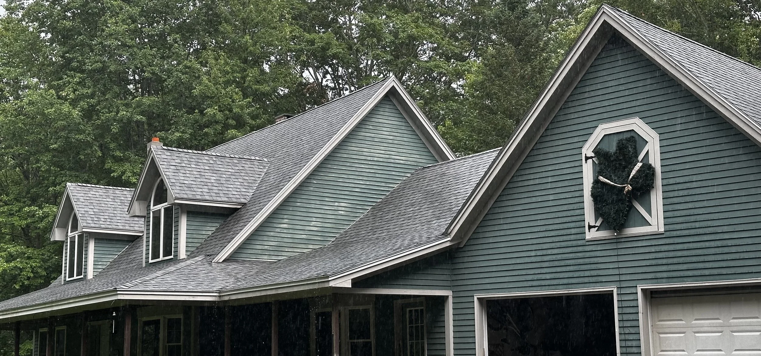 A house with teal siding and gray shingle roof, featuring three small dormer windows and a large gable with a window decorated with a black heart-shaped wreath.