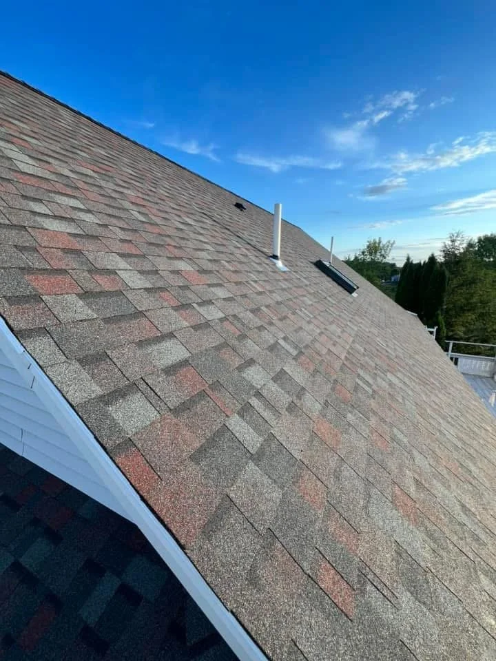 View of a sloped roof with asphalt shingles in shades of gray and red, featuring two vents and a skylight, with a background of blue sky and some clouds.