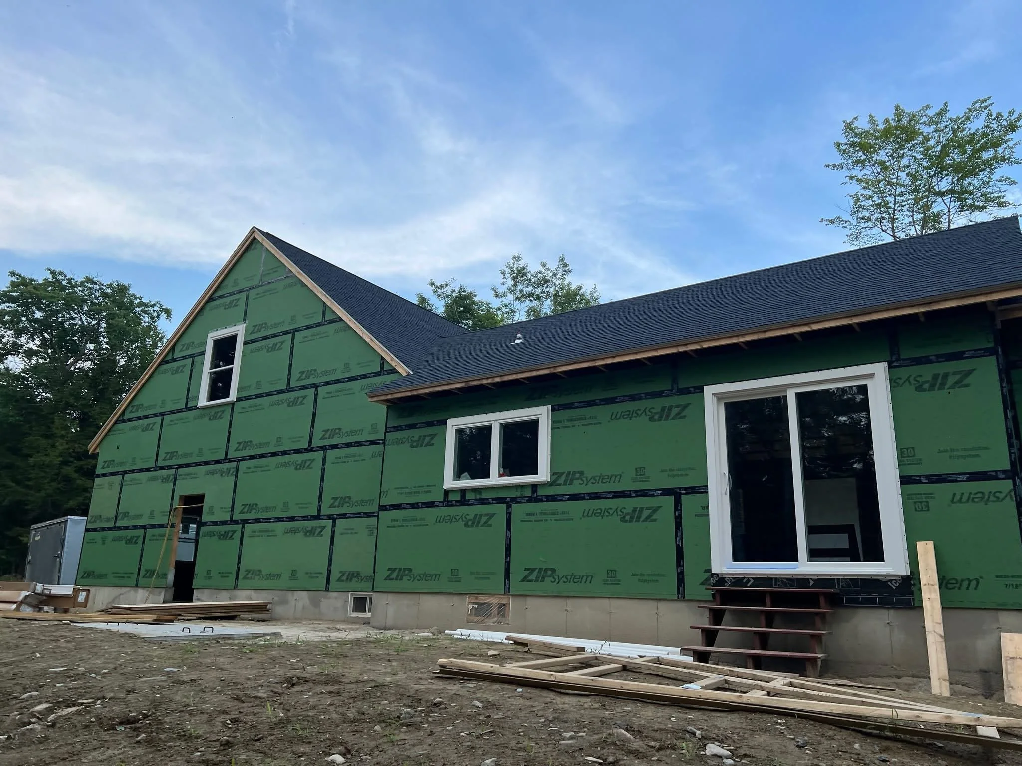 Construction site of a house with green exterior sheathing, white window frames, and a dark roof; scaffolding and building materials are on the ground.
