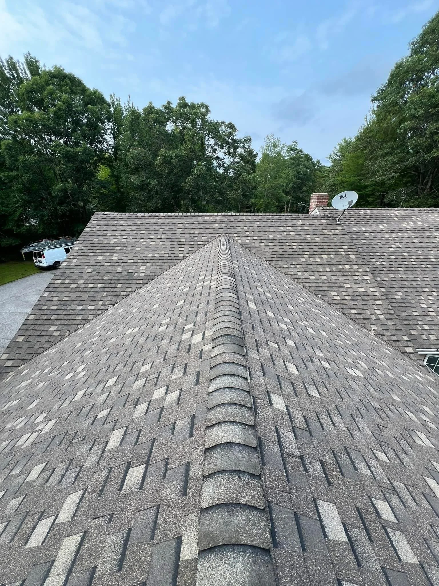 A view of a residential roof with asphalt shingles, showing the ridge and side slopes, bordered by green trees and a partly cloudy sky.