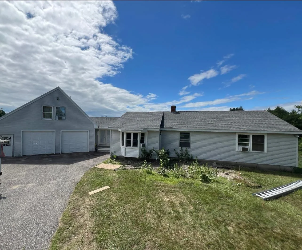 Front view of a white house with attached garage, small plants in front, and a ladder on the lawn under a partly cloudy sky.