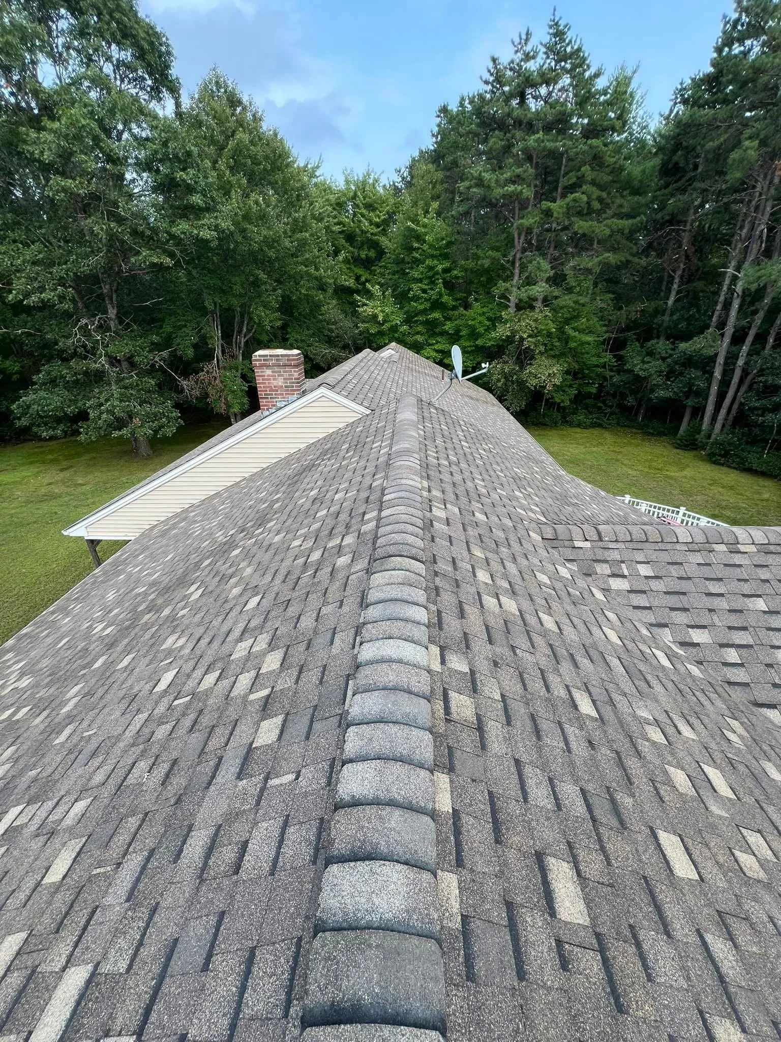 View looking down a sloped shingled roof of a house with a chimney and a satellite dish, surrounded by green trees and a cloudy sky.