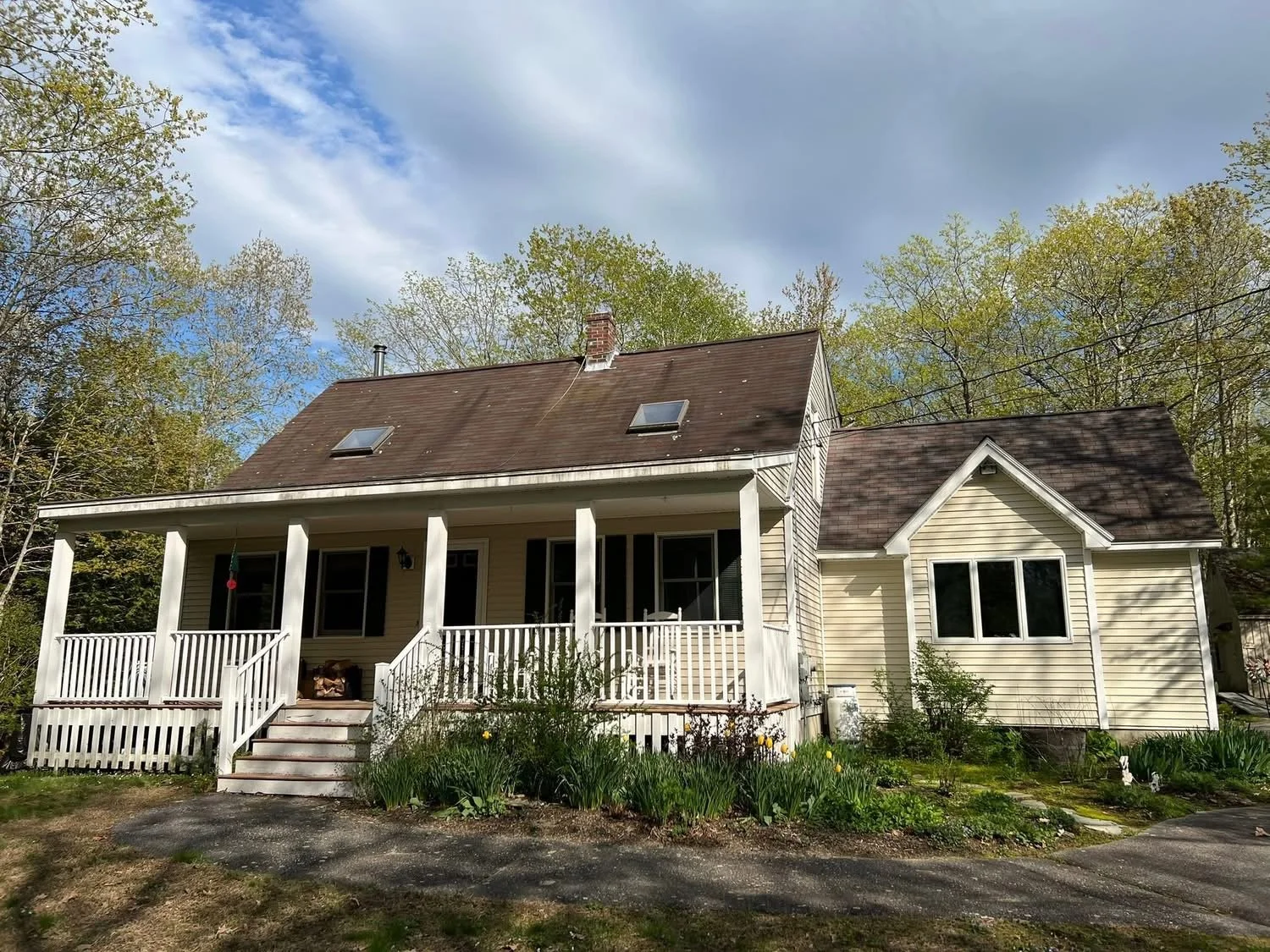 A two-story house with a porch and a gabled extension. The house has a brown roof and light-colored siding. The porch has white railings and steps leading up to it. There are two skylights on the main roof and several trees in the background.