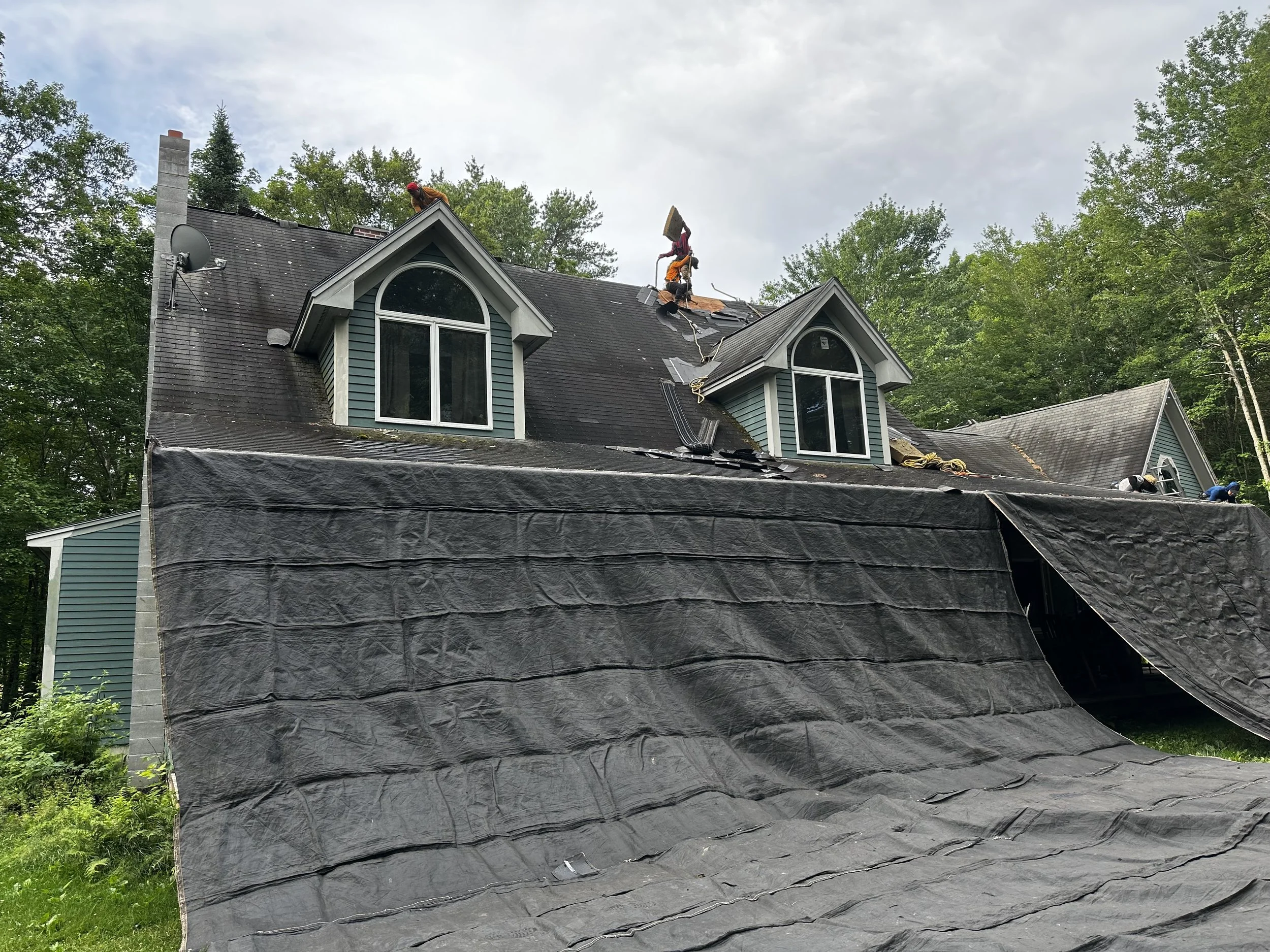 Roofing workers are repairing the roof of a house during the daytime. Some parts of the roof are open, with roofing materials and tools nearby, while other parts are covered with a tarp.