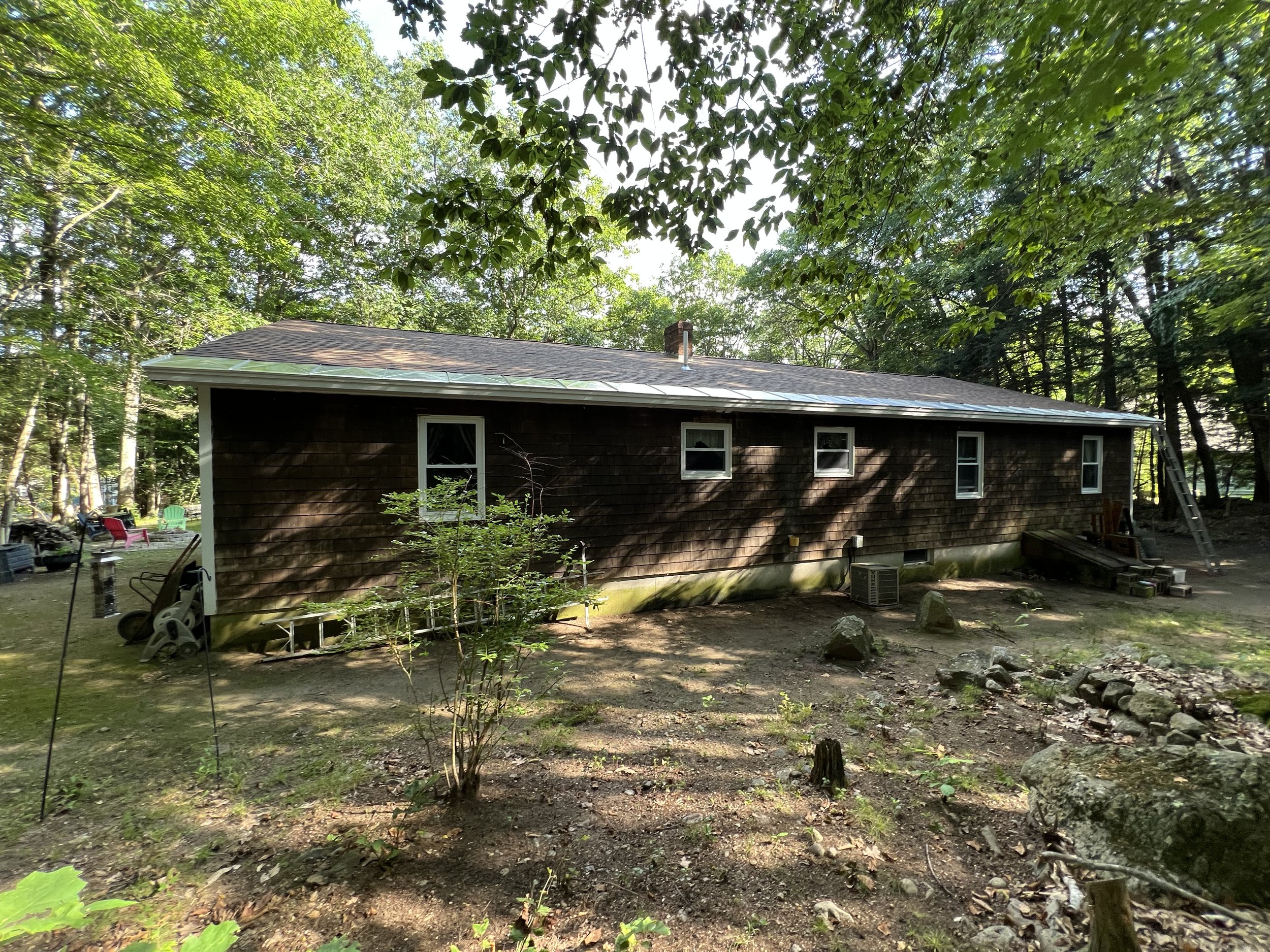 Back view of a brown house with four windows, surrounded by trees and rocks on the ground, with outdoor furniture and equipment nearby.