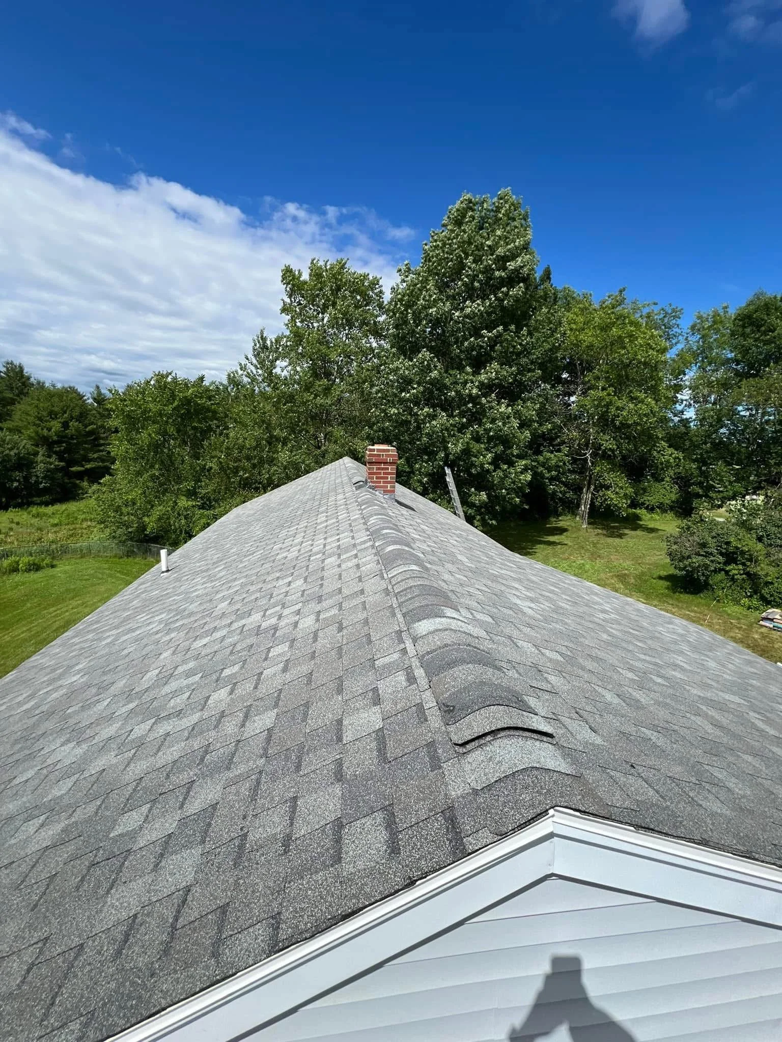 View of a house roof with gray shingles, a brick chimney, and surrounding green trees under a partly cloudy blue sky.