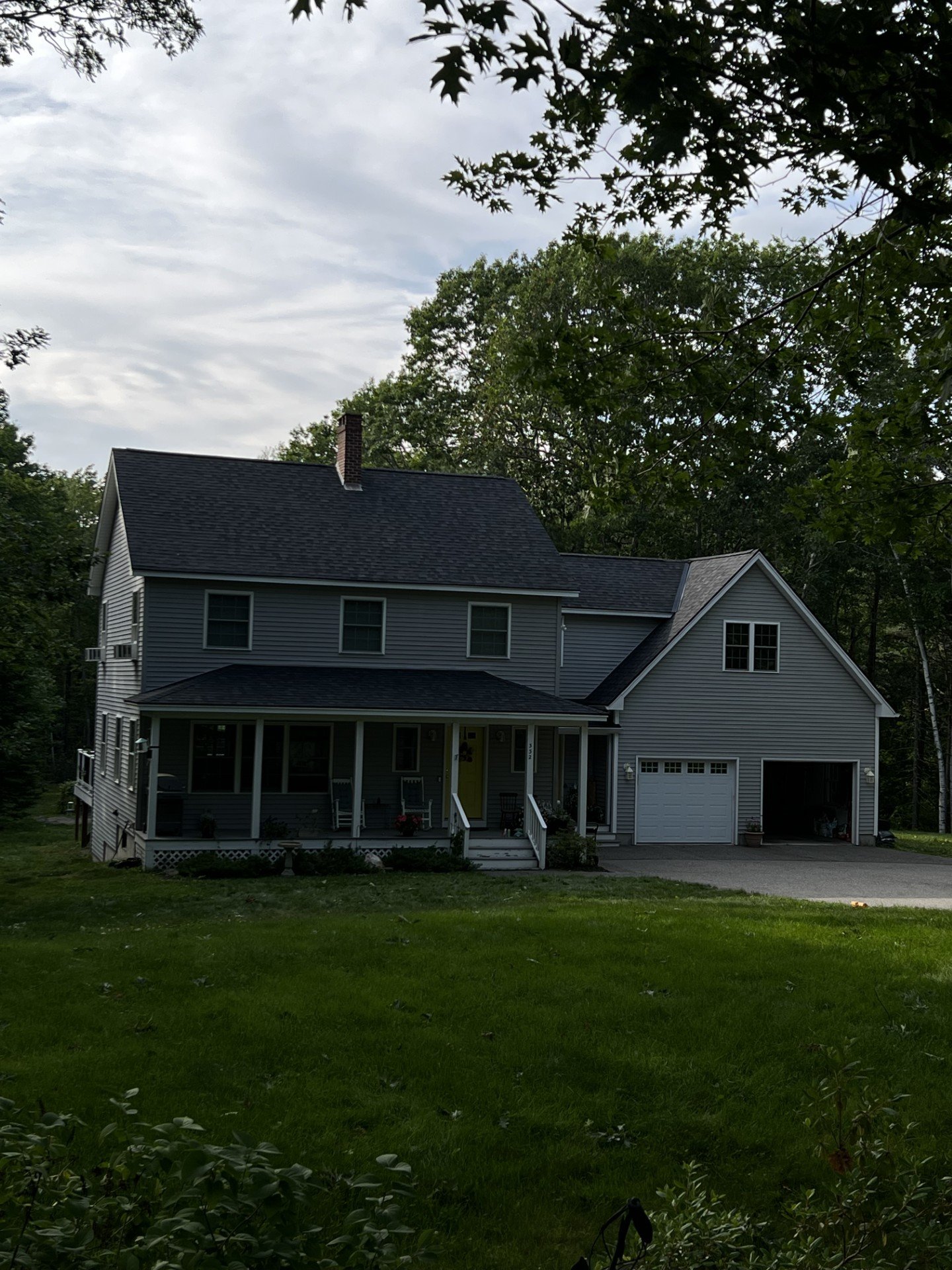 A two-story gray house with a front porch, a one-car garage, and a green lawn. Tall trees surround the house under a partly cloudy sky.