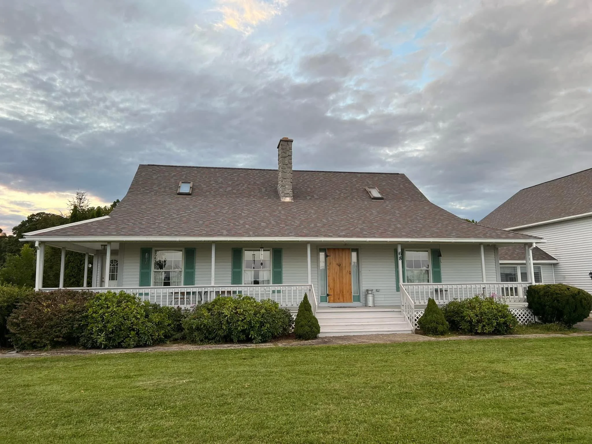 A house with white siding, green shutters, and a brown front door, surrounded by bushes and a grassy lawn, under a cloudy sky.