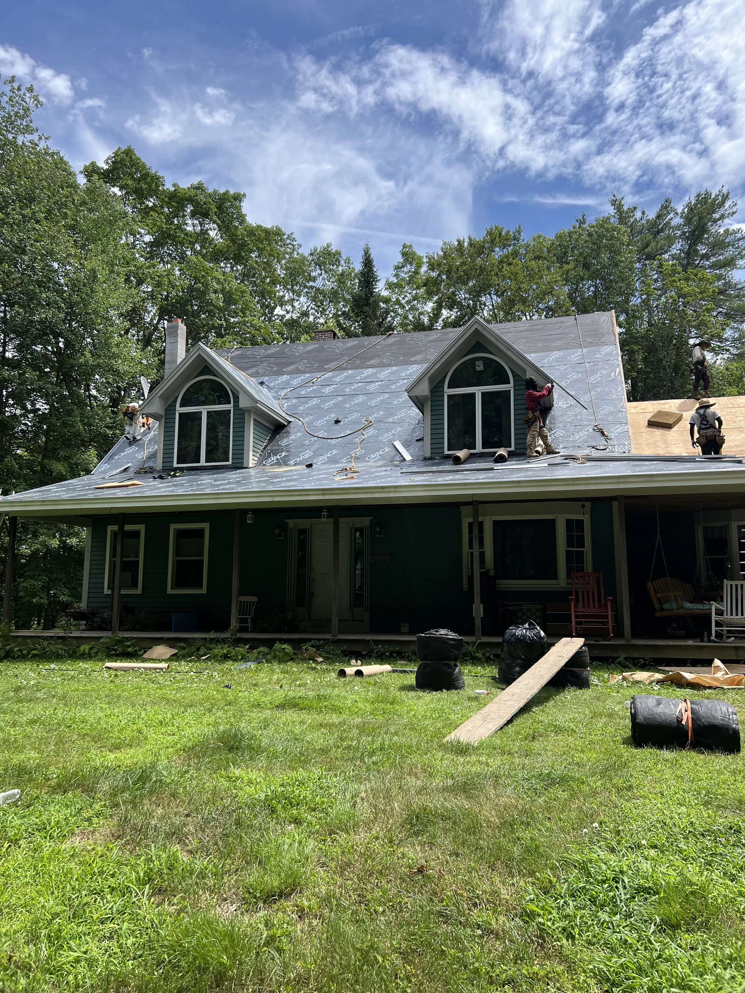 Construction workers installing new roofing on a house with a green exterior, two dormer windows, and a porch with outdoor furniture, under a partly cloudy sky.