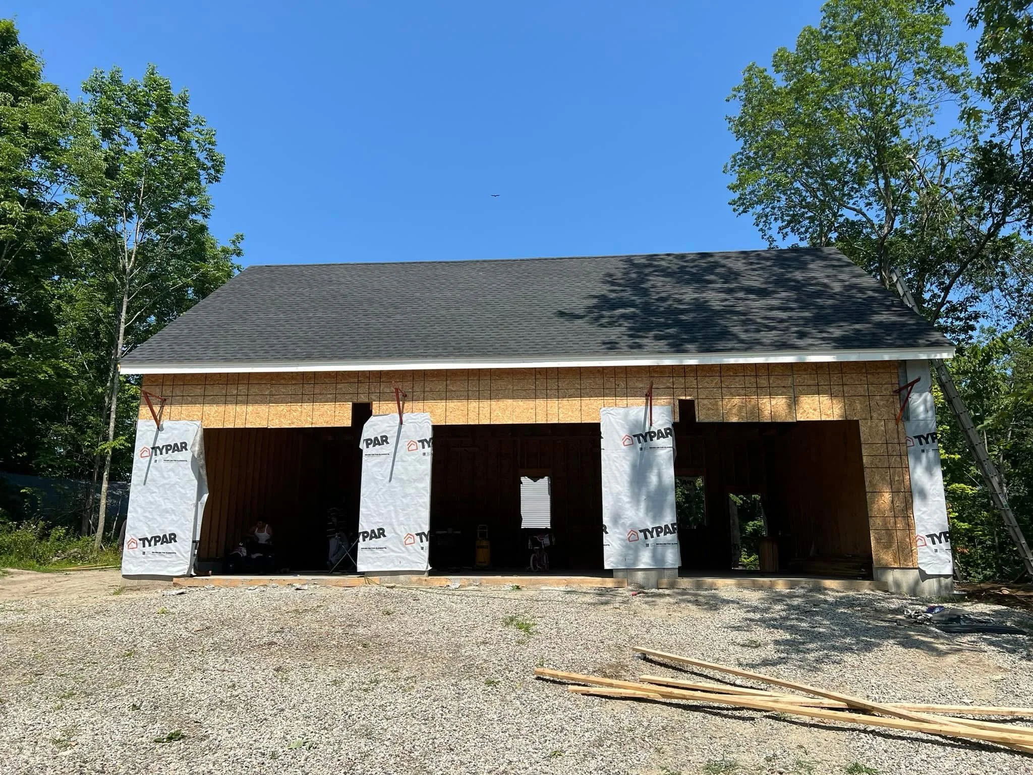 Construction of a house with a dark grey roof and partly finished exterior walls, surrounded by trees and a gravel ground, with construction materials on the ground.