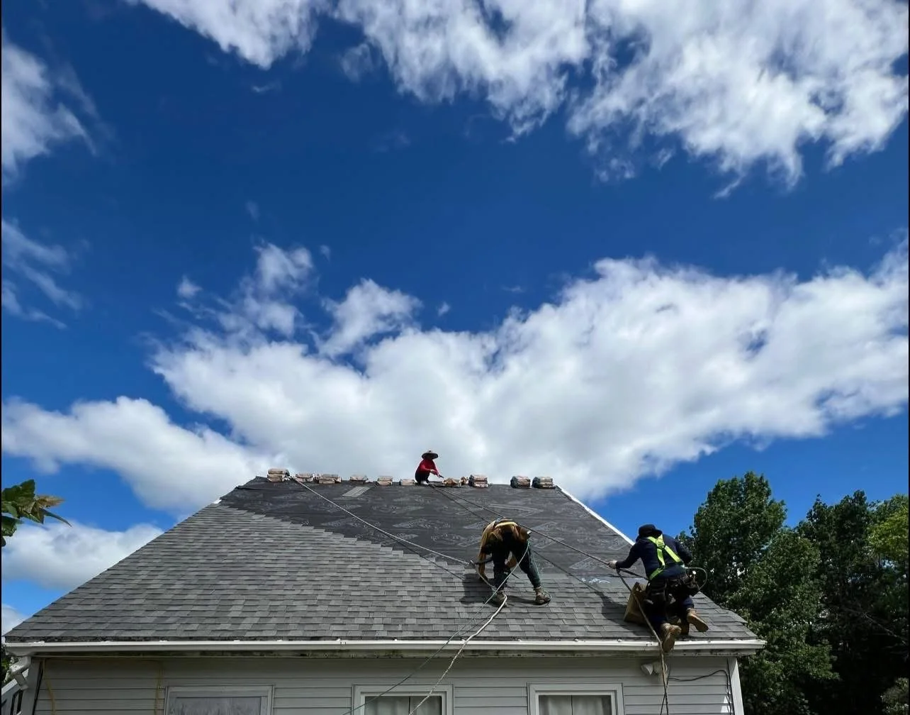 Three workers repairing or installing a roof on a residential house, with blue sky and clouds in the background.