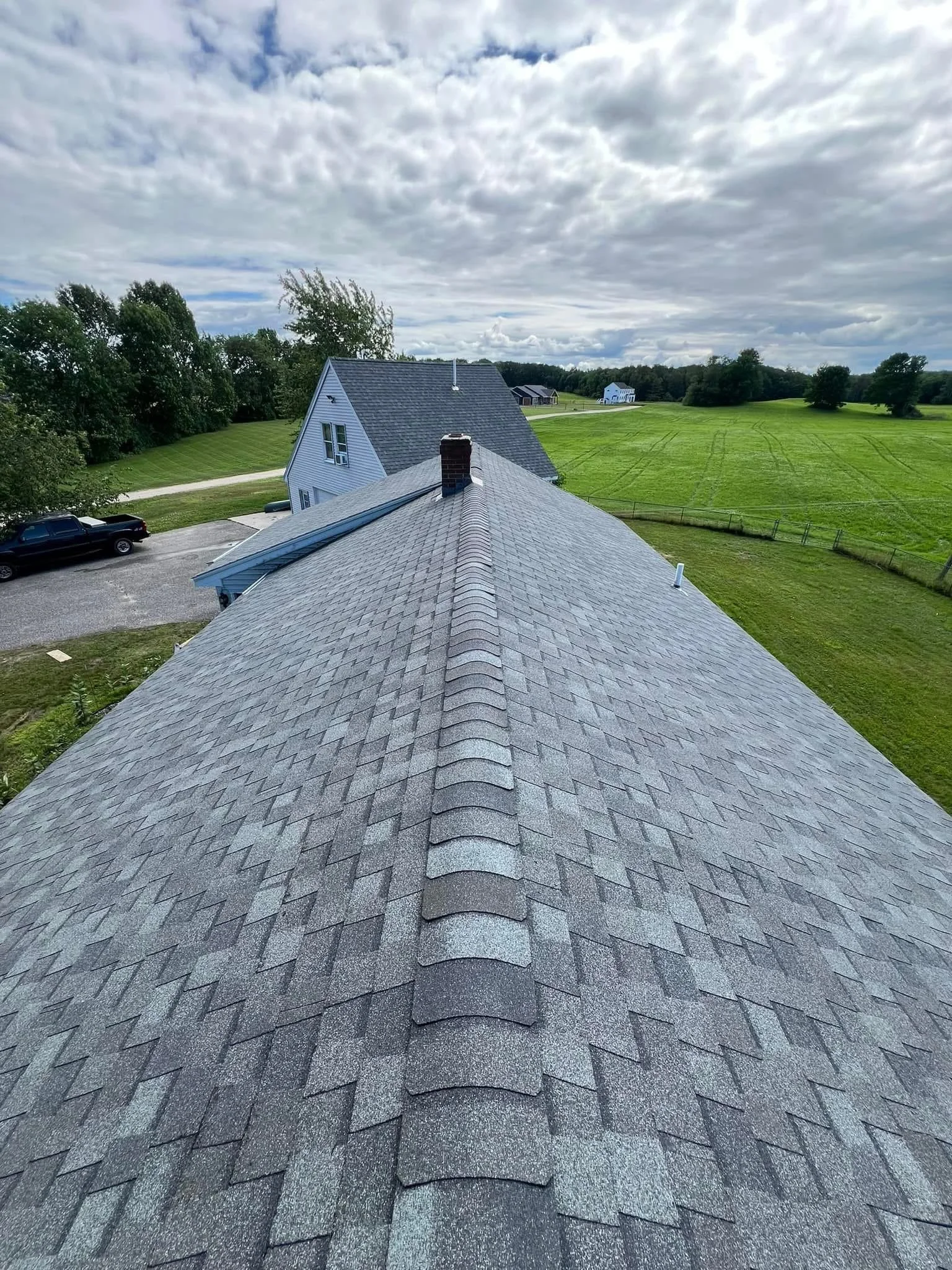 View from the roof showing asphalt shingles, a chimney, and the surrounding landscape with other houses, trees, and a cloudy sky.