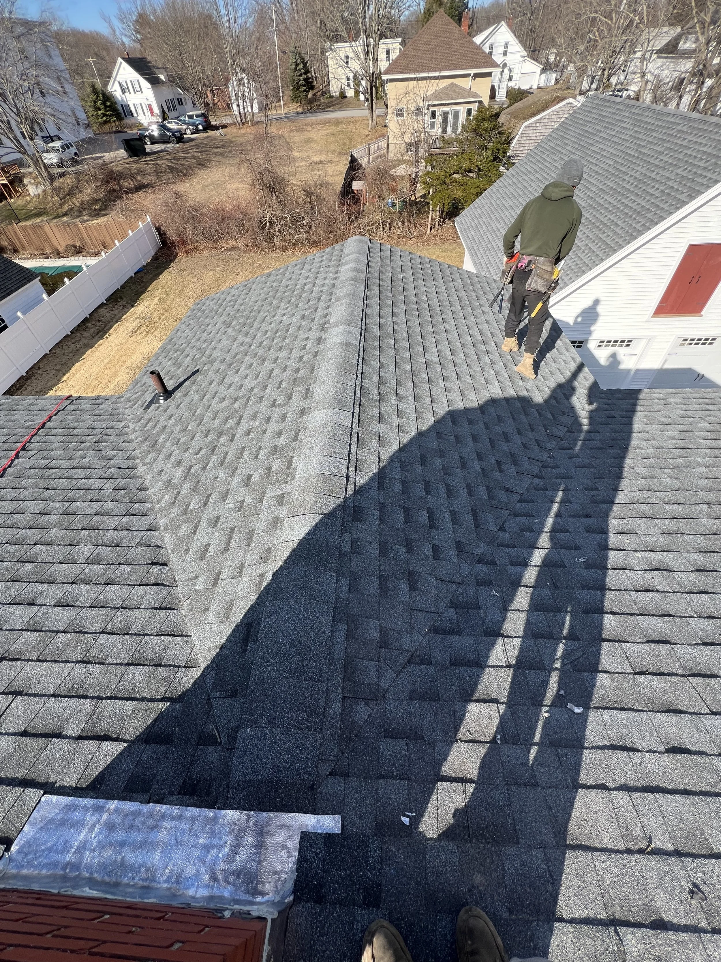 A person standing on a rooftop in a residential neighborhood, wearing work clothes and a tool belt, with a shadow cast on the roof. The neighborhood has multiple houses, trees, and parked cars.